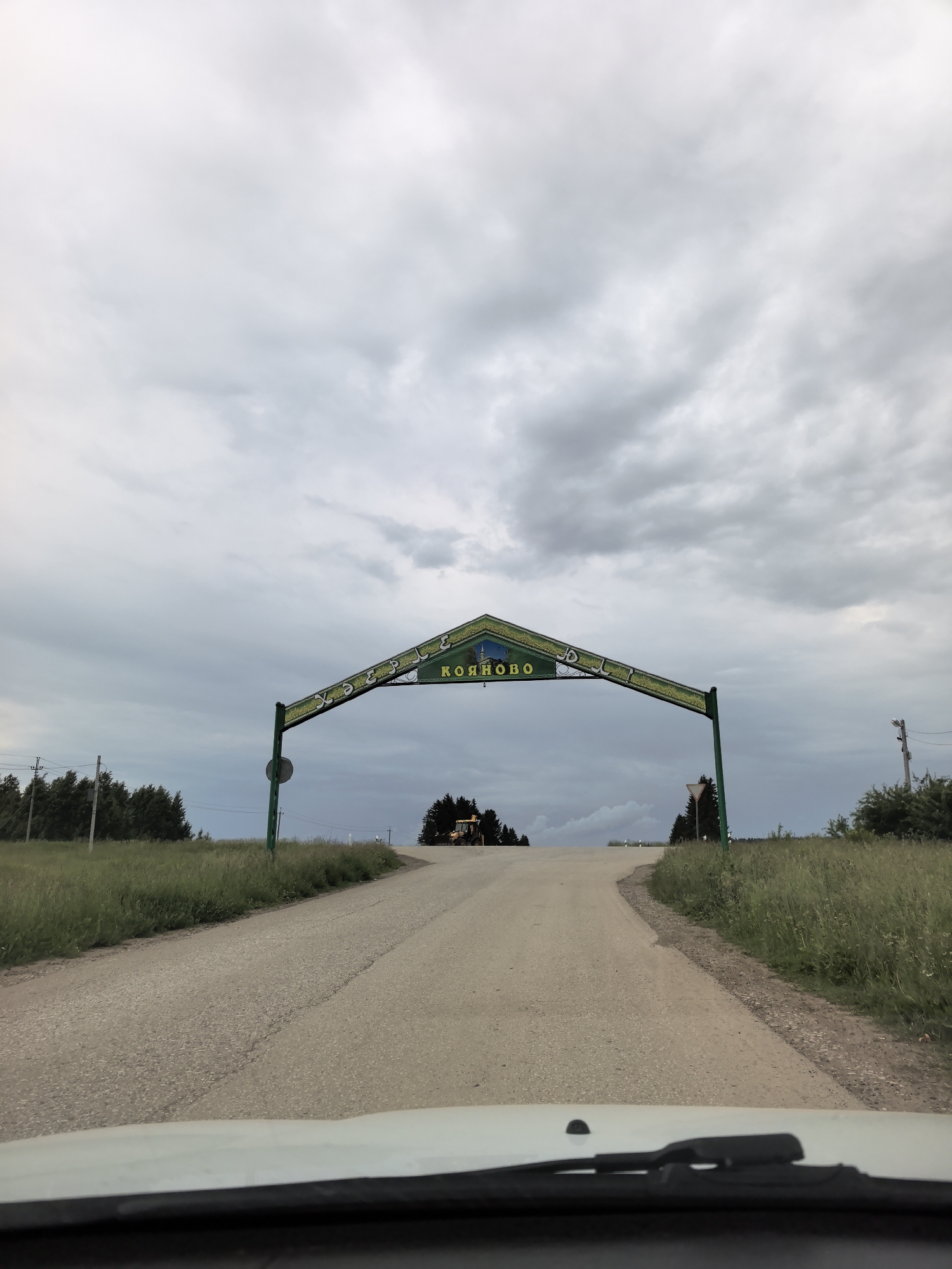 The image shows an overhead archway sign with the Russian text "КОЯНОВО" in yellow letters against a green background. The arch spans a paved road that curves to the right. The sky is overcast with gray clouds. On the left side of the road, there is a field of tall green grass and a line of trees. On the right side, there is also grass and trees, and further in the distance, a yellow bulldozer is visible. The photograph appears to be taken from the perspective of someone inside a vehicle, looking forward at the sign.
