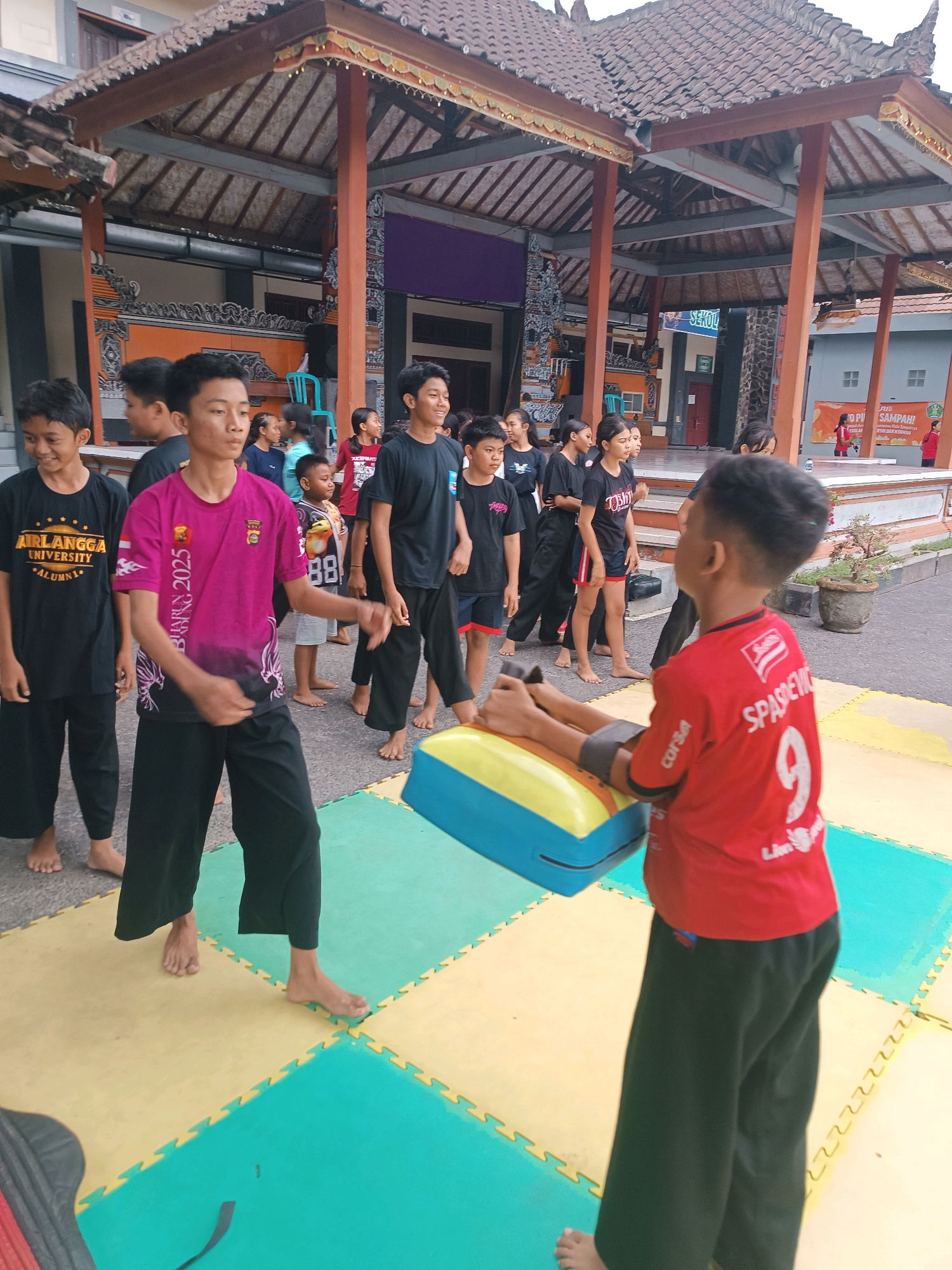 In Selemadeg, Indonesia, a group of young people, likely students, are engaged in an outdoor training session on a mat area. The scene is set in front of a traditional Indonesian building with a pitched roof supported by wooden pillars.
In the foreground, two boys are the primary subjects. The boy on the left, wearing a pink t-shirt with "BHARU 2025" printed on it and black pants, is in motion, with his arms outstretched and his body turned to the right. He appears to be demonstrating a move or practicing. To his right, another boy wearing a red t-shirt with the number "9" and "SPASHEVIC" printed on the back, is positioned behind a blue and yellow training pad, which is resting on the ground. His arms are extended towards the pad, suggesting he is either holding it or has just struck it.
Behind these two boys, a line of other young individuals, both male and female, are observing. They are dressed in casual attire, mostly black t-shirts and dark pants. Some are looking towards the central action, while others seem to be waiting for their turn.
The ground is covered with a mat composed of interlocking yellow and green puzzle pieces. In the background, the traditional building features intricate carvings and decorative elements, characteristic of Balinese architecture. The overall atmosphere appears to be one of focused activity and communal learning within a cultural setting.