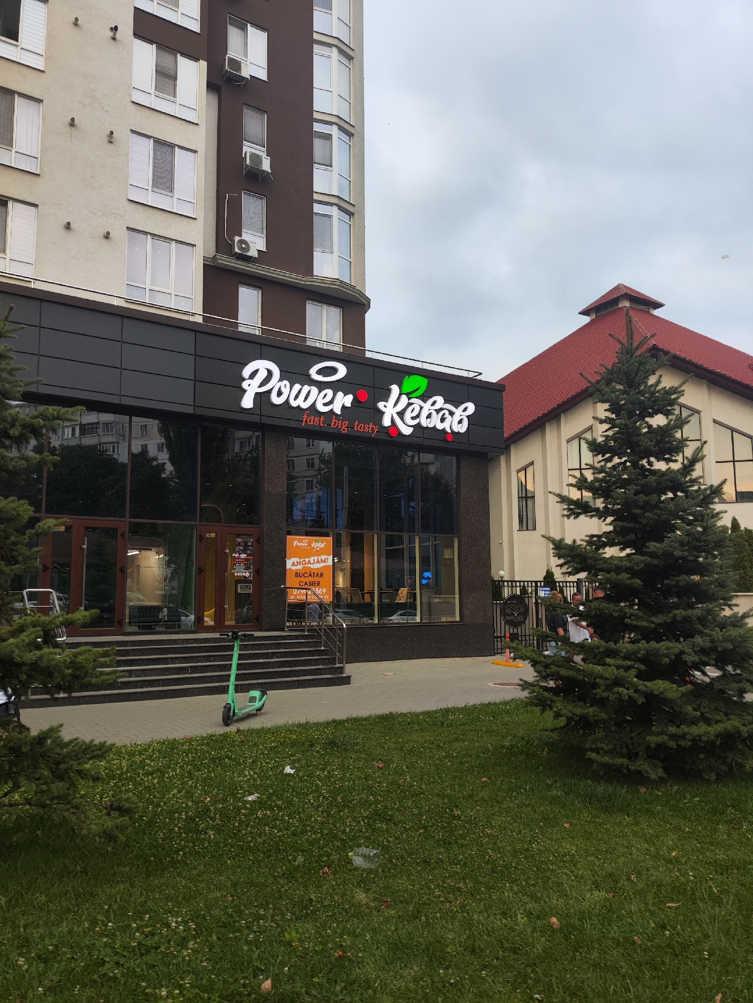 A medium shot captures the exterior of a restaurant with prominent signage reading "Power Kebab". The restaurant appears to be a ground-floor establishment within a multi-story building. To the left, a section of the building features a lighter beige facade with multiple windows and air conditioning units. To the right of the restaurant, a separate building with a distinctive red, pitched roof is visible. In front of the restaurant, a set of stairs leads to a glass-fronted entrance. A green electric scooter is parked on the paved area in front of the stairs. A patch of green grass with scattered fallen leaves is in the foreground. The sky is overcast. The Power Kebab restaurant is located in Chișinău, Moldova.