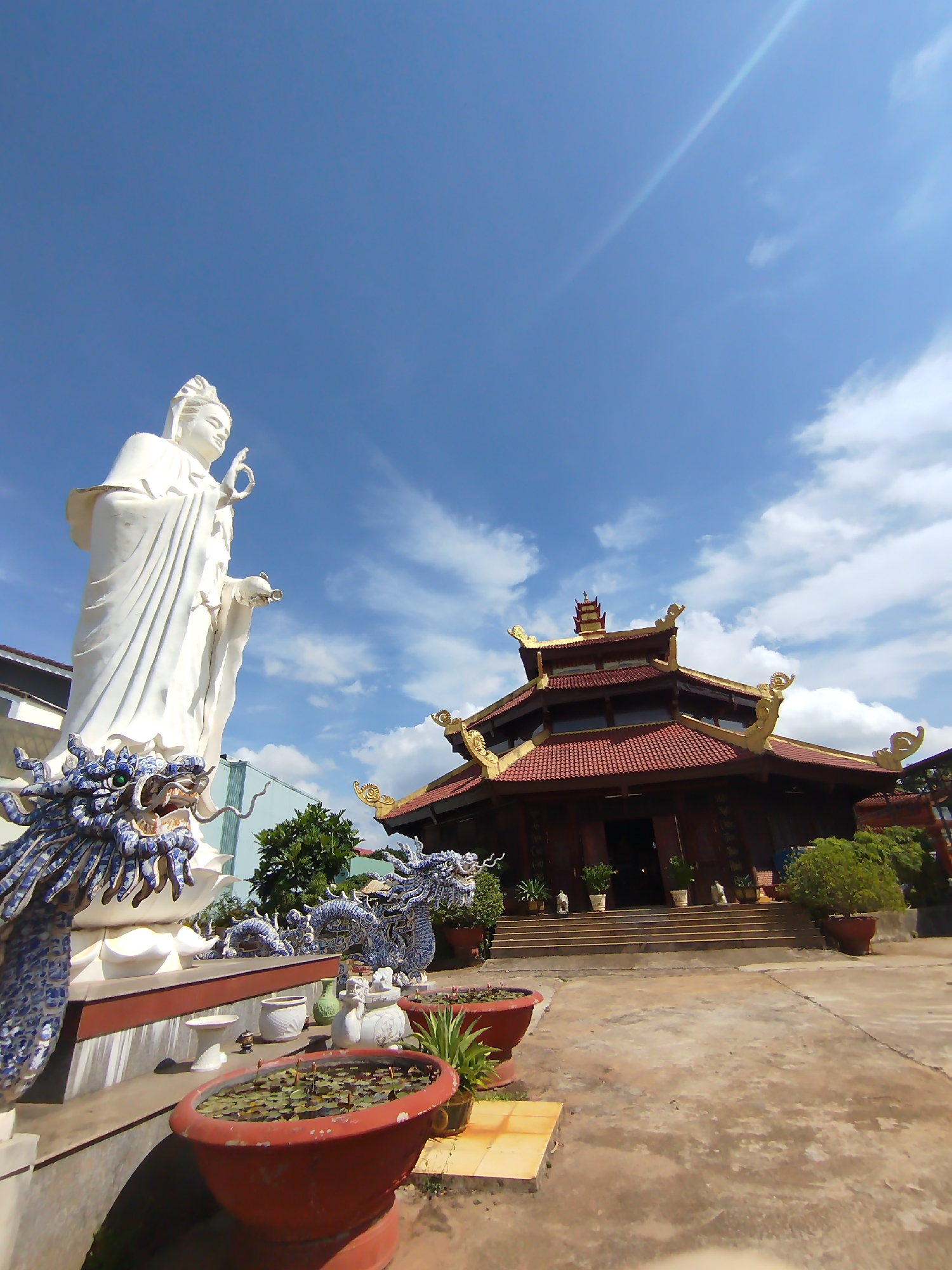 The image shows a courtyard in Pleiku Ward, Vietnam. Dominating the left side is a large white statue of a Bodhisattva, adorned with a blue and white dragon motif at its base. Several potted plants, some containing lotus flowers, are arranged around the statue. To the right stands a traditional Vietnamese-style building with a red tile roof and golden ornamentation. The sky above is blue with scattered clouds. The courtyard is paved with concrete. No people are visible in the frame.