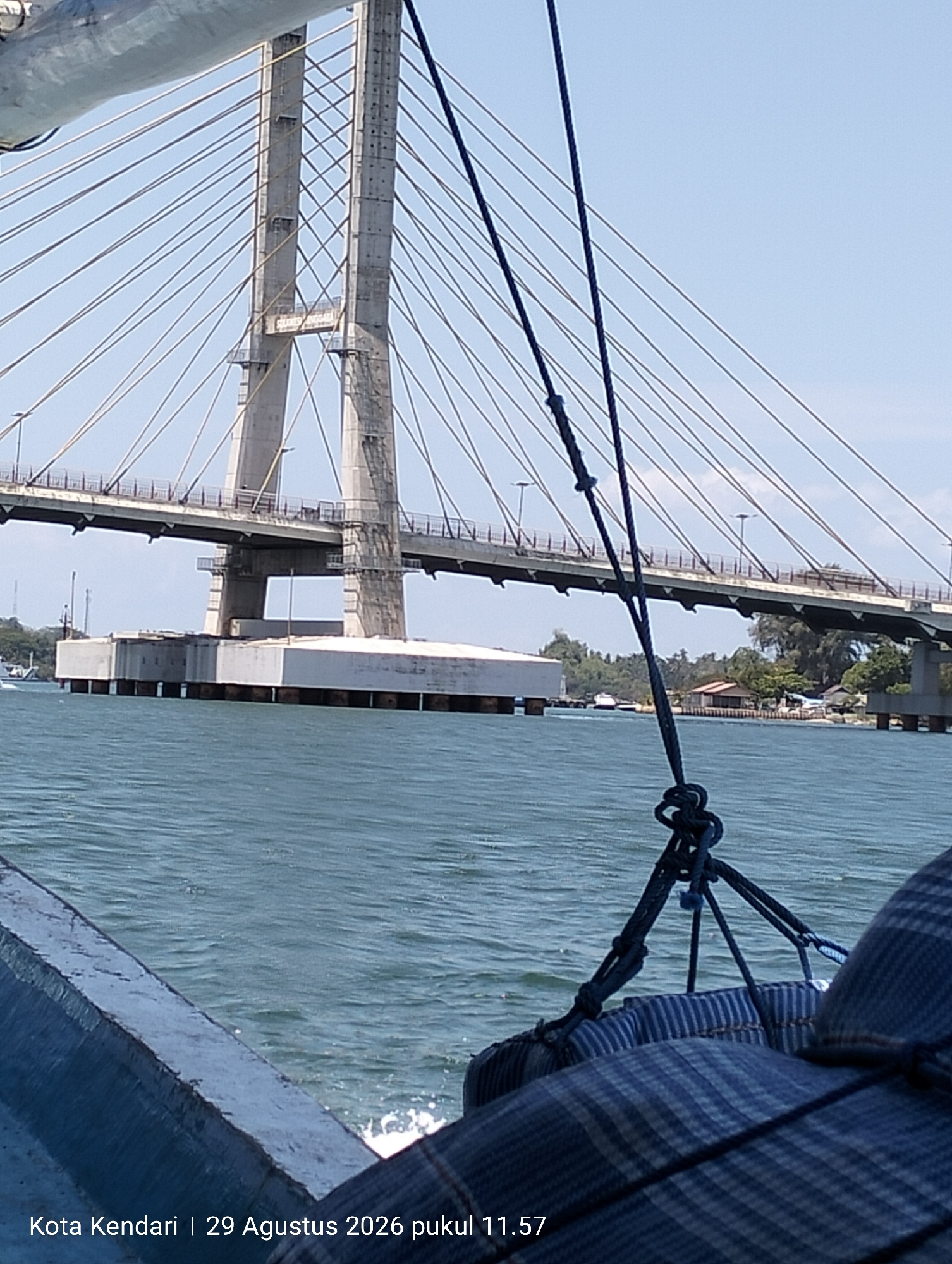 The image shows a view from a boat in Kendari, Indonesia, on August 29, 2026, at 11:57 AM. The main subject is a large cable-stayed bridge, likely the Kendari Bay Bridge, spanning across a body of water. The bridge's concrete pylons and numerous cables are visible against a clear blue sky. In the foreground, the edge of the boat is visible, with a blue upholstered seat and some dark ropes tied with nautical knots. The water is a deep blue-green, with some small waves. In the background, the opposite shore is lined with trees and some buildings, including what appears to be a small house on stilts. A white boat is visible on the water near the shore. The scene is under bright daylight.