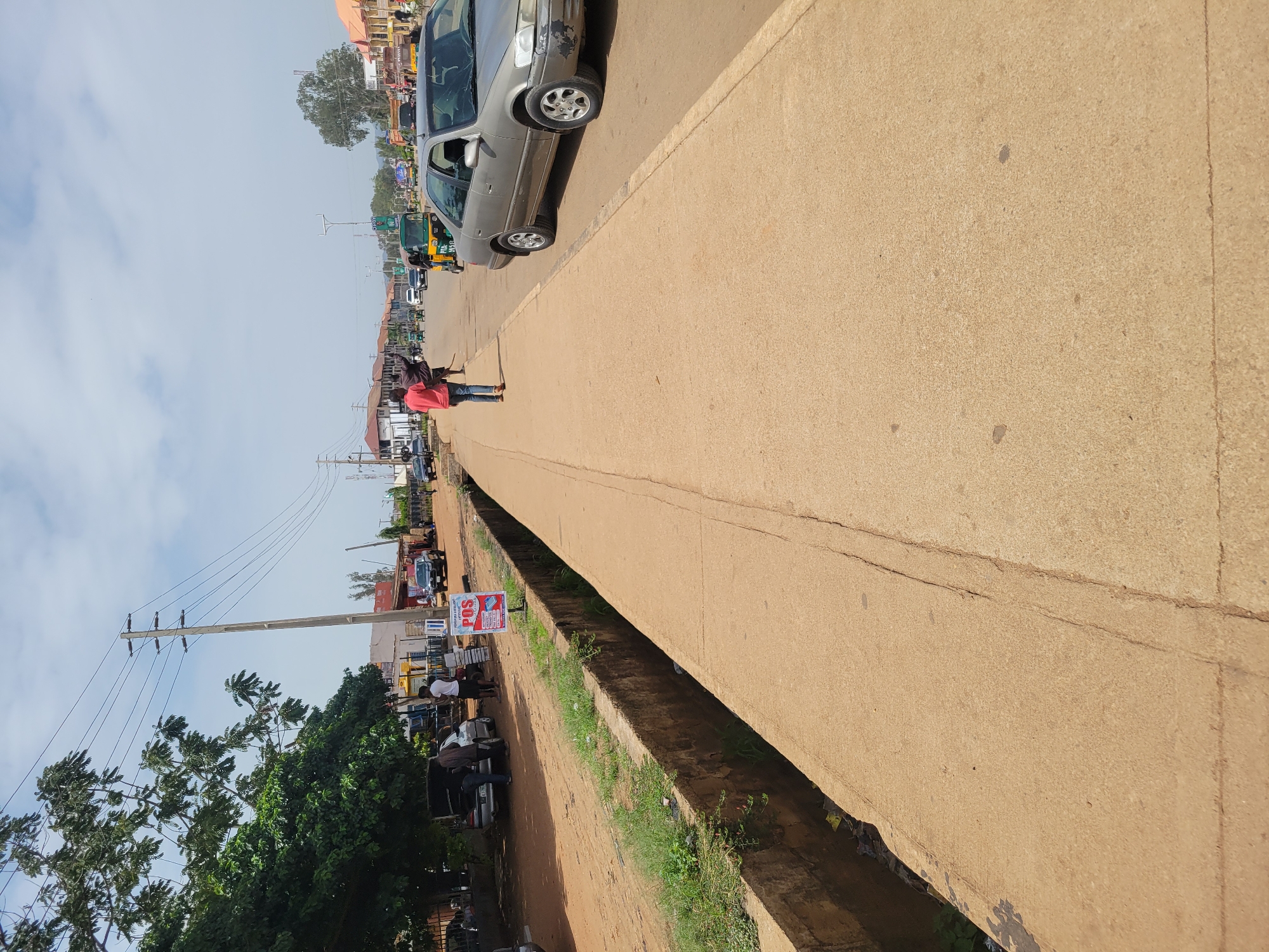 The image shows a street in an urban area. In the foreground, there is a concrete structure, likely a bridge or overpass, with a drainage ditch running alongside it. The street features various vehicles, including cars and motorbikes. A person in a red top is standing in the middle of the road. There are buildings visible on both sides of the street, including shops and residences. In the lower left corner, there is a tree and a utility pole with power lines. A sign labeled "POS" is visible next to people who look to be making a transaction. The sky is overcast.