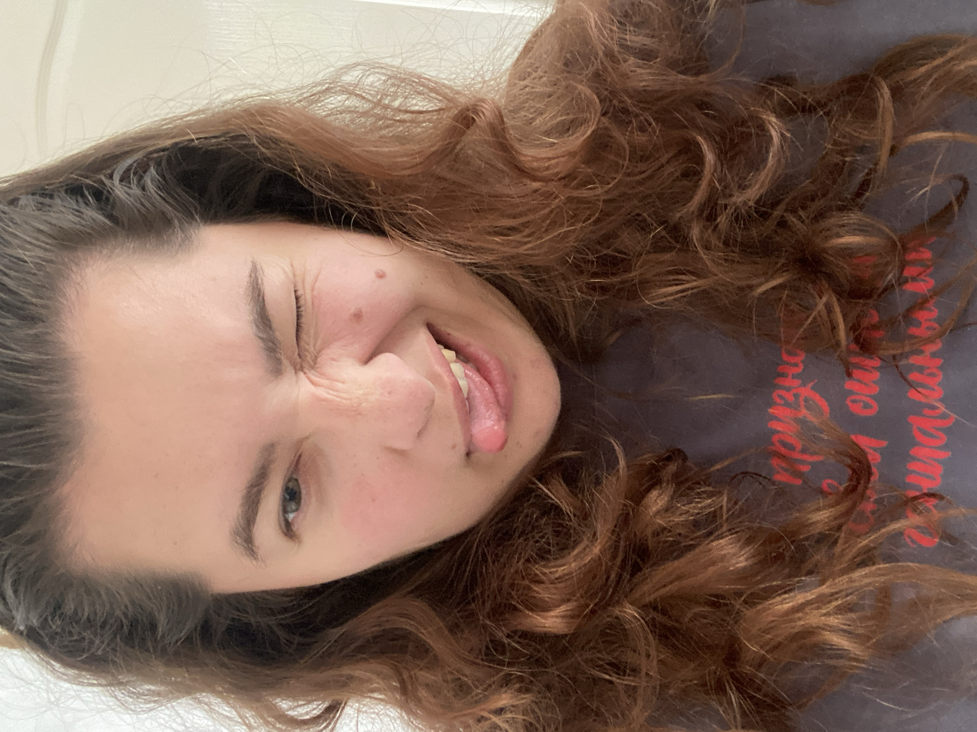 A medium shot captures a young woman with auburn, wavy hair. She is making a face, winking with her right eye and sticking out her tongue. Her left eye is open and looking at the camera. She is wearing a dark gray t-shirt with red lettering. The background is out of focus and appears to be an indoor setting, possibly an apartment. There is no indication of the city of Perm, Russia, in the image.