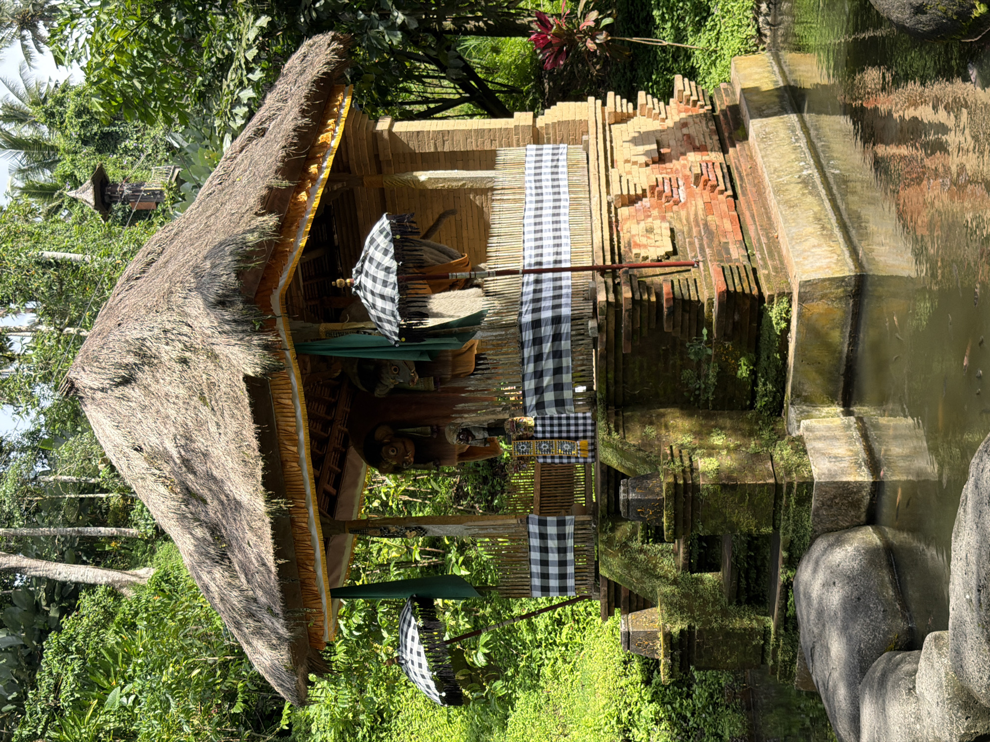 The image depicts a small, thatched-roof structure, possibly a shrine or pavilion, located at Tegal Suci, Indonesia. The structure features brick and stone elements, with a checkered black and white cloth draped across part of its facade. A black and white umbrella is also present. Lush green vegetation surrounds the structure, and a body of water reflects part of it. The scene suggests a tranquil and possibly sacred space within a natural environment.