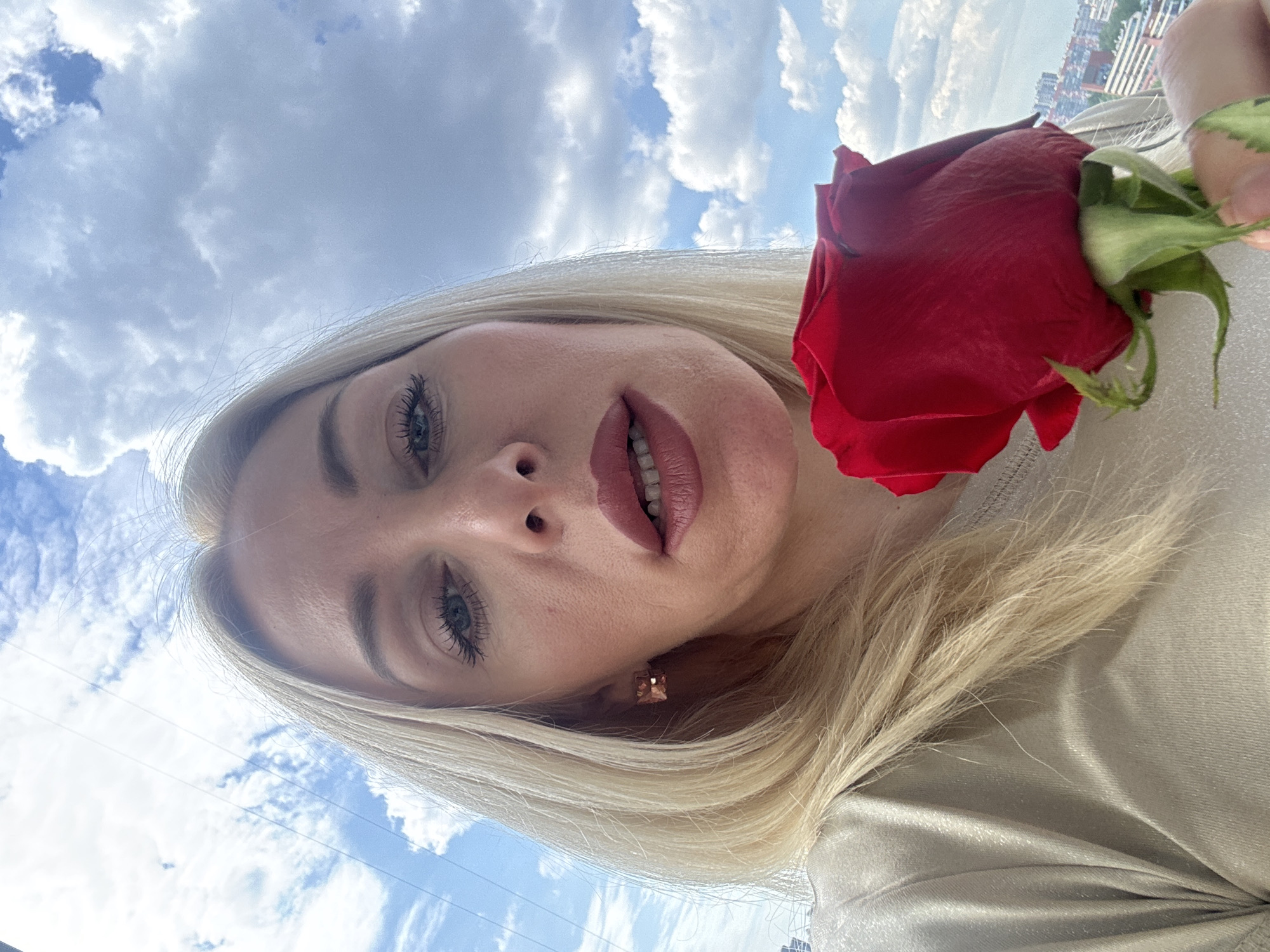 A close-up selfie captures a blonde woman looking directly at the camera. The image is oriented with the woman's head towards the left side of the frame and her body extending to the right, suggesting it was taken while looking upwards or was rotated. She has light blue or grey eyes with visible mascara and eyeliner, and her lips are painted with a soft pink or nude lipstick. A square, possibly rose-gold, earring is visible in her right ear.
She holds a vibrant red rose with green sepals and a visible stem near her right shoulder. Her long, light blonde hair frames her face and drapes over her shoulder. She appears to be wearing a light-colored, possibly champagne or silver, garment with a smooth, slightly reflective texture.
The background is predominantly a bright blue sky filled with numerous fluffy white clouds. In the far distance, towards what would be the bottom right of an upright frame, a blurry cityscape with several buildings is visible, indicating a high vantage point, possibly from a balcony or rooftop. The scene depicts daytime with fair weather. There is no visible text in the image.