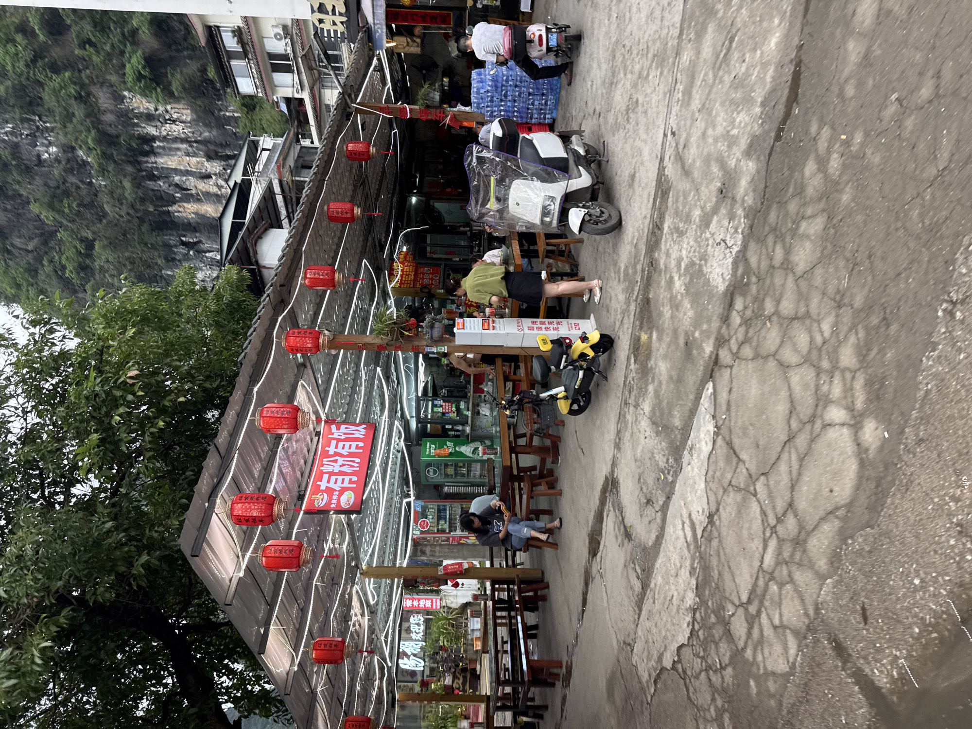 Here's a detailed description of the image:
**Scene:** The image shows a street scene with a restaurant in Guilin, China. The restaurant's entrance is adorned with red lanterns and illuminated signs in Chinese characters.
**People:** There are several people in the image. One woman in a green shirt and black skirt appears to be interacting near the counter of the restaurant. Another person in the back appears to be loading water jugs onto the back of his scooter. A young person is seated and looking at his phone.
**Objects:** The scene features various objects, including:
* Electric scooters parked near the restaurant.
* Red lanterns hanging from the restaurant structure.
* Tables and wooden stools arranged in front of the restaurant.
* Signage with Chinese characters advertising the restaurant and the food and beverages they offer (e.g., rice noodles and local dishes).
* Refrigerators and drinks visible inside the restaurant.
* A partially visible mountain slope in the background.
**Actions/Interactions:** People are either interacting with the restaurant (possibly ordering or paying) or seated and occupied with their phones. Others appear to be setting up scooters with water jugs.
**Environment:** The area seems to be an open-air setting, possibly a street market or a roadside eatery. The pavement appears wet or damp. The background includes what appears to be an apartment building next to the mountain.