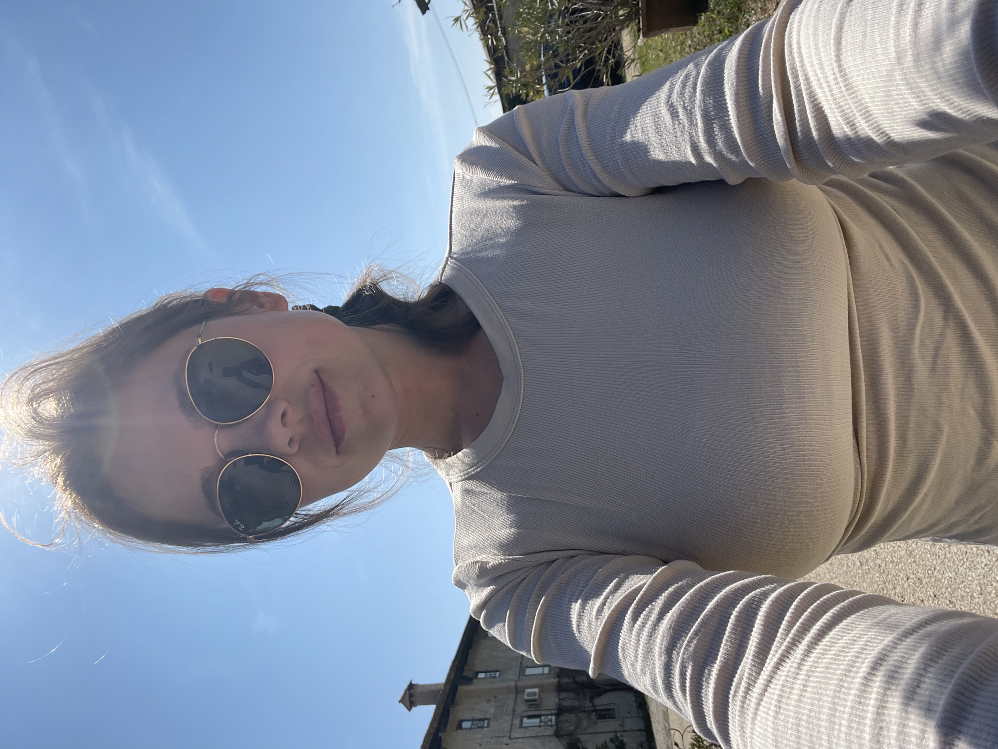 A woman wearing sunglasses and a long-sleeved ribbed shirt is taking a selfie on a sunny day in San Lorenzo, Italy. The sky is clear blue. In the background, there's a building with windows and a small tower-like structure. The ground appears to be paved.