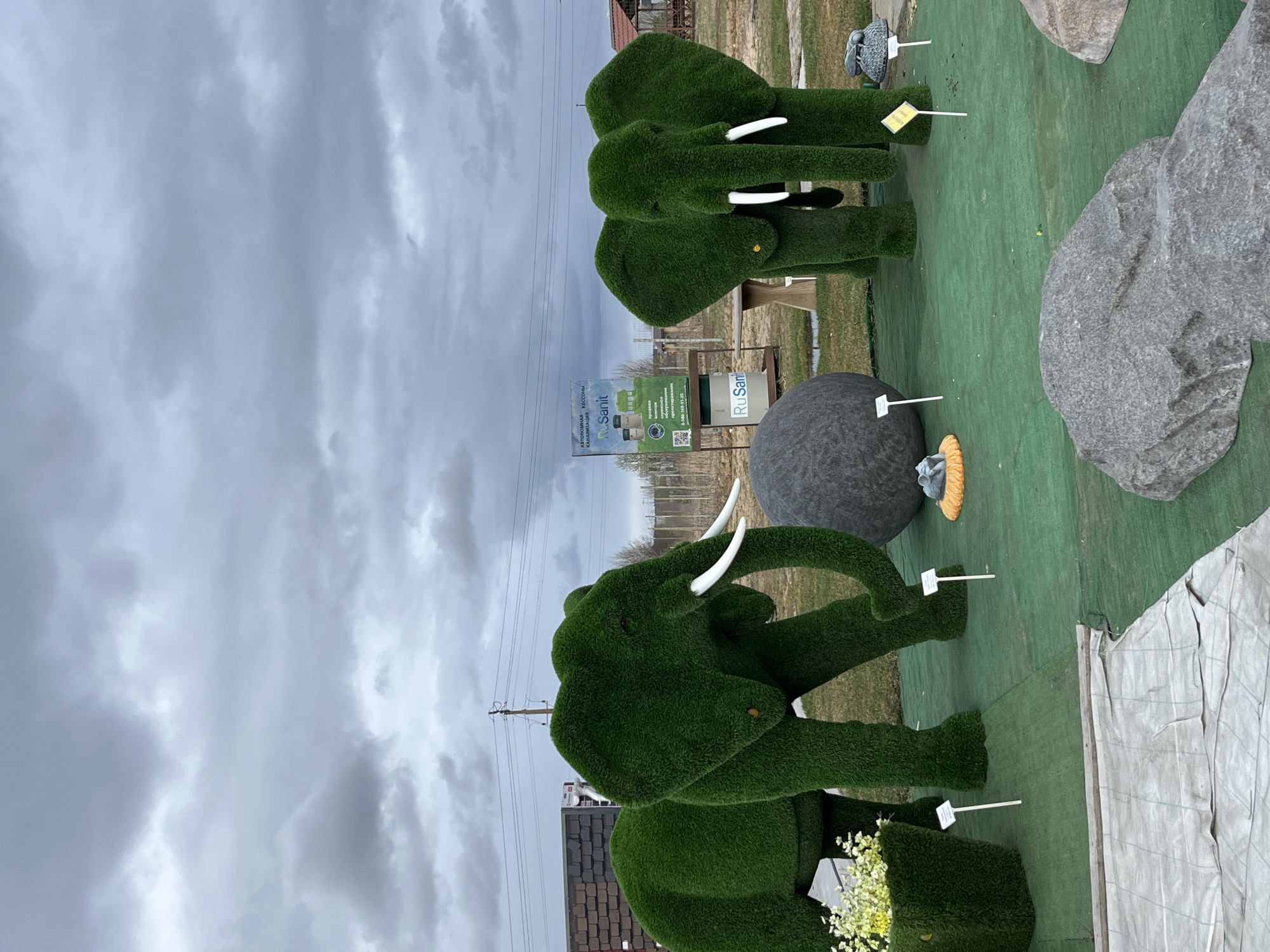 The image shows a display of green, moss-covered elephant sculptures and decorative stones in an outdoor setting, likely a sales area, in Bezborodovo, Russia. Two elephant sculptures are the primary focus; one is larger and facing the viewer, and the other is smaller and situated further back. They both have prominent white tusks. The area around the sculptures is covered with artificial green turf. Between and behind the elephants are several gray rocks or stones. In the background, there is a sign with the words "RuSanit" in Cyrillic lettering and a QR code. The sky above is overcast and gray. A partial view of a building and power lines are also visible in the background.