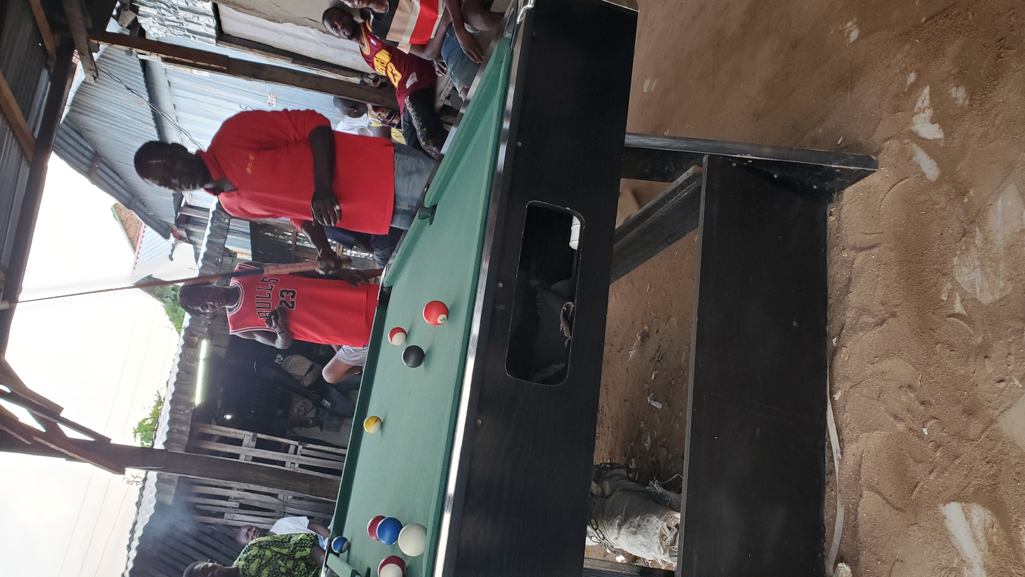 In Jalingo, Nigeria, a group of men are gathered around a pool table outdoors, under a makeshift shelter. The pool table is green with black padding and is covered in various colored billiard balls, including red, white, blue, and yellow. Two men in red shirts are actively engaged with the pool table, one appearing to hold a cue stick. Several other men are visible in the background, some seated and observing the game. The environment suggests an informal setting, with a dirt ground and simple wooden structures forming the shelter. The overall scene depicts a casual social gathering centered around a game of pool.
