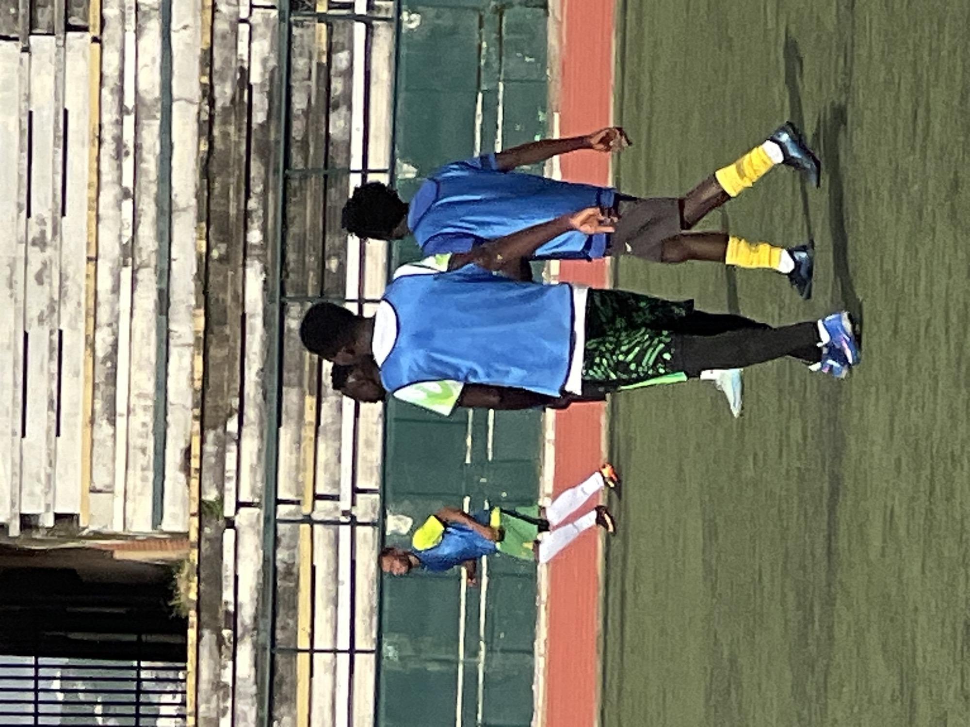 The image depicts a group of young men playing soccer on a grassy field at the New Stadium in Nigeria. The stadium seating, visible in the background, is characterized by layered concrete and metal structures. The players are dressed in athletic wear, including blue vests over t-shirts and shorts. One player, in the foreground, is wearing green shorts with a black and green pattern, black leggings, and blue and white soccer cleats. Another player in the mid-ground is wearing green shorts, white socks, and orange and black soccer cleats. The field itself is green artificial turf with a red track bordering it. The overall lighting suggests it is daytime, possibly late afternoon, with shadows indicating the sun's position. The scene captures a moment of athletic activity within an urban stadium setting.