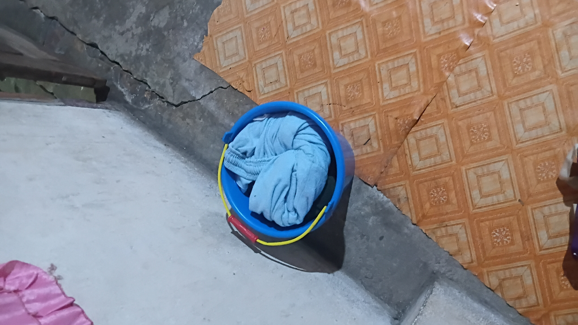 A blue plastic bucket containing folded light blue clothing is situated on a cracked, tiled floor. The bucket has a yellow handle with a red grip. To the left of the bucket, a white concrete wall is visible. In the upper left corner, a portion of a pink, ruffled fabric is draped over the wall. The scene appears to be indoors, with a dark, possibly wooden structure in the background. The overall impression is that of a domestic setting, possibly a utility area or a room under renovation, located in Prabumulih, Indonesia.