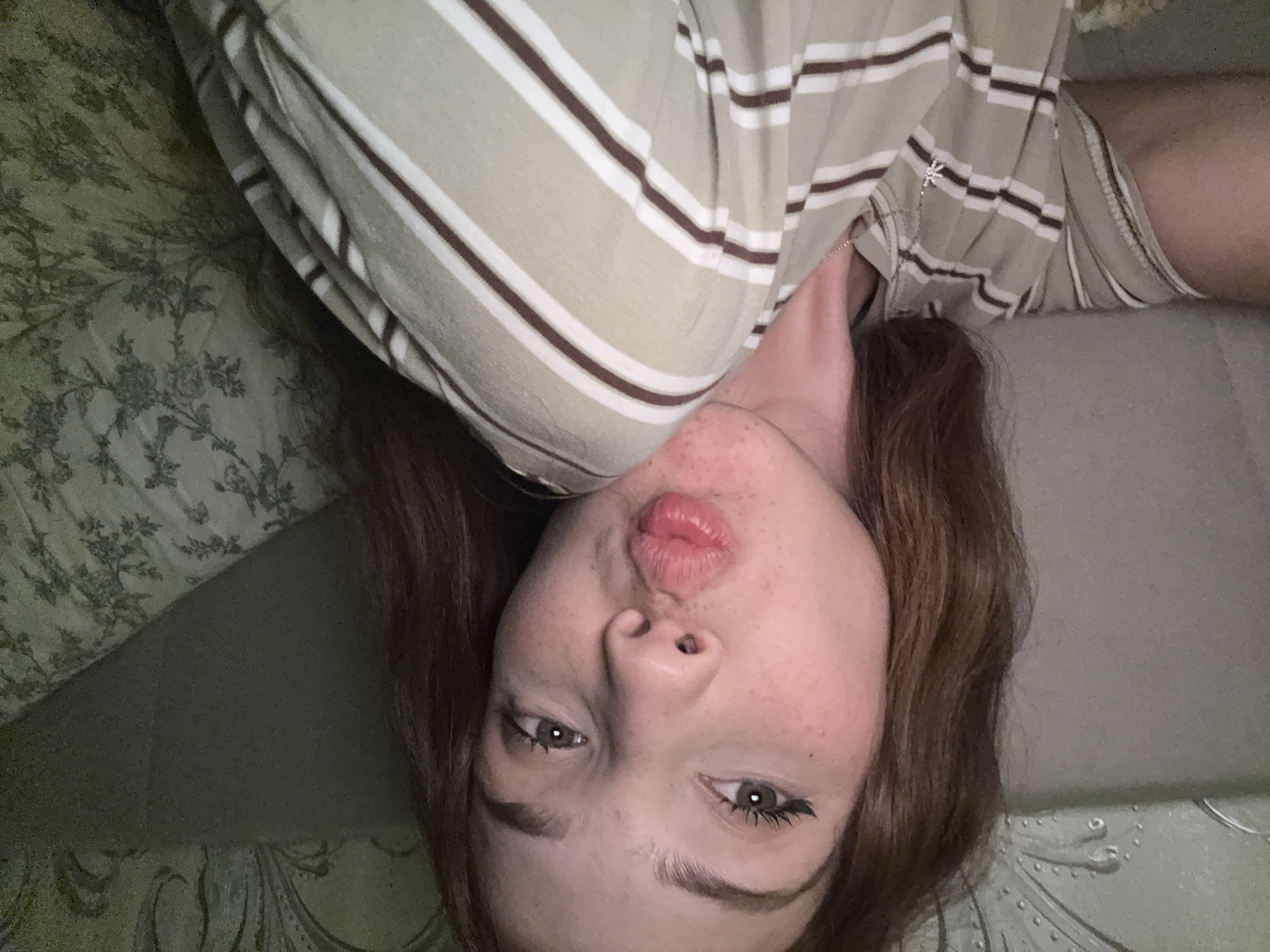 The image is a close-up, upside-down selfie of a young person with auburn hair, lying down on a textured surface. They are wearing a light-colored t-shirt with horizontal brown and white stripes. The background is a muted green with a subtle floral pattern on the left side and a plain gray surface on the right. The person is pouting, with their lips pursed. Their eyes are open, and they have dark eyeliner on their upper lashes. Small reddish marks are visible on their cheeks, possibly acne. A silver necklace with a small, star-shaped pendant is visible at the neckline of their shirt. The lighting appears to be dim, suggesting it might be nighttime or indoors. The scene is intimate and personal, with the subject directly engaging the camera. There is no visible text in the image.