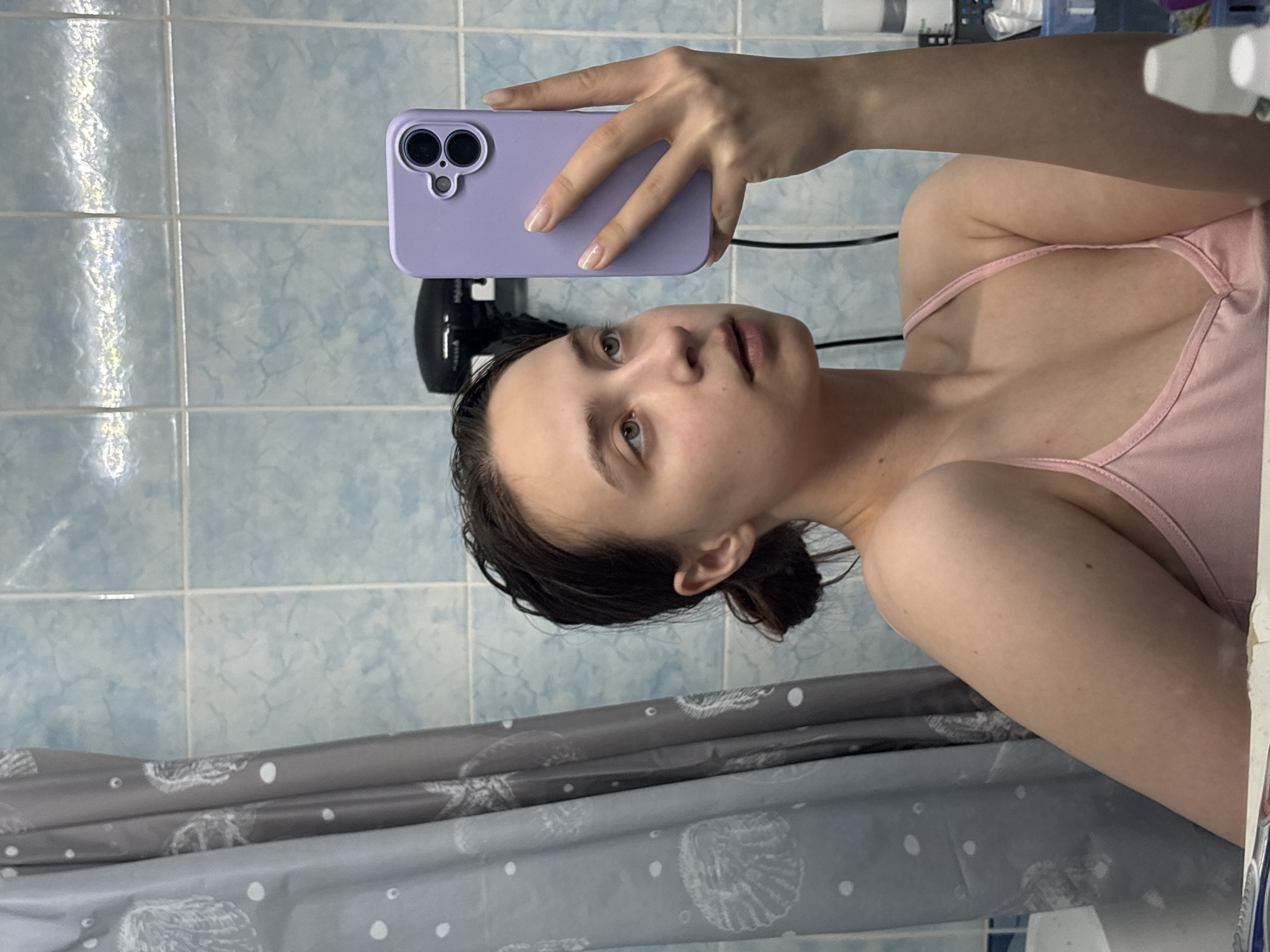 A young woman is taking a selfie in a bathroom. She's wearing a light pink tank top and holding a purple phone case. She has wet dark hair pulled back in a bun. The bathroom has light blue tiled walls and a gray shower curtain with white shell patterns. A black hairdryer is mounted on the wall behind her. The location is likely in Прикамский, Russia.