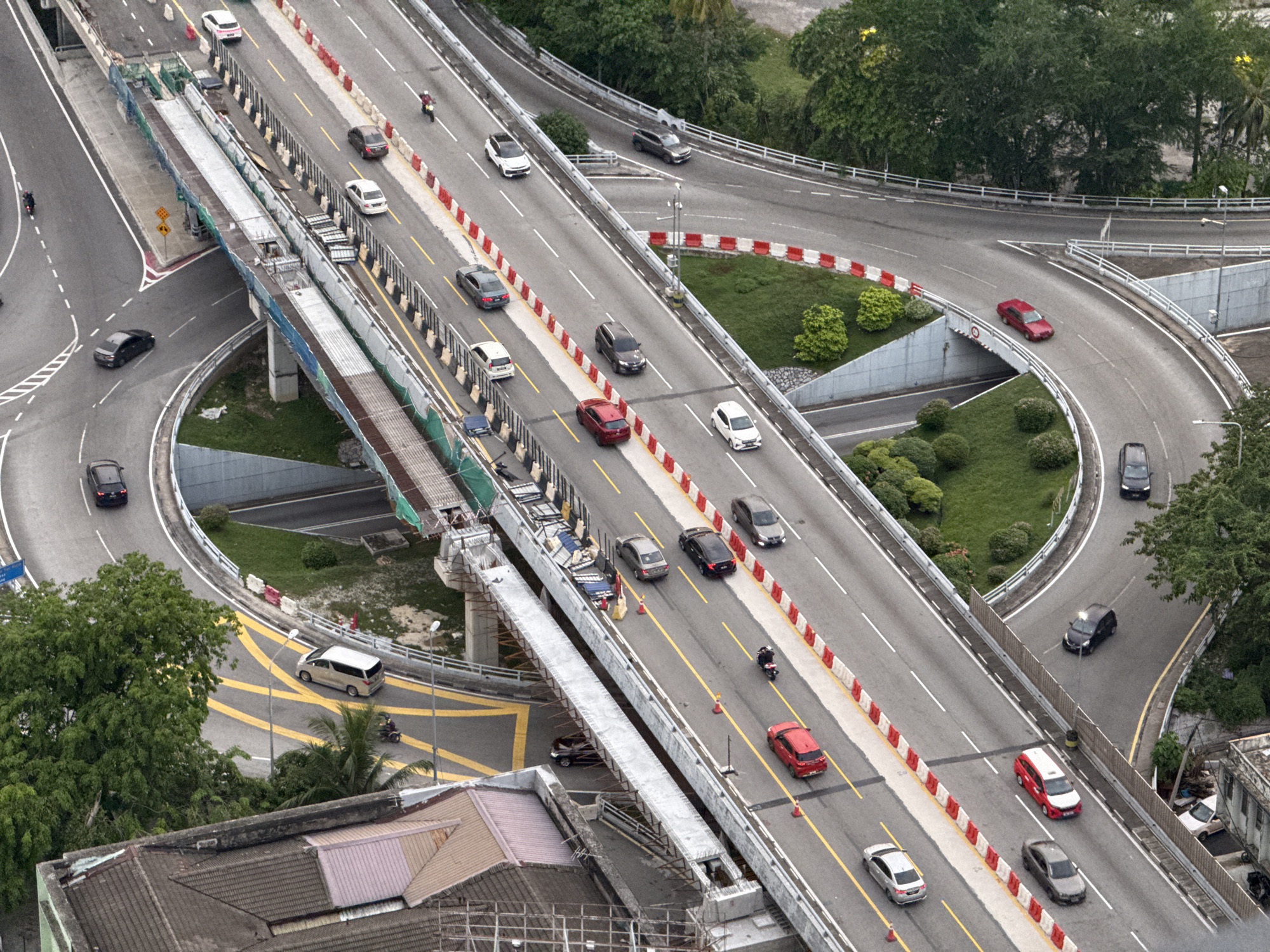 This aerial photograph depicts a section of a highway interchange in Kuala Lumpur, Malaysia. Multiple roadways intersect and branch off, facilitating traffic flow. The primary roadway runs horizontally through the frame, with guardrails separating the lanes. Numerous vehicles, including cars and motorcycles, travel along the roads. An elevated construction site spans part of the highway, marked by scaffolding and construction materials. Adjacent to the roadways are landscaped areas with greenery. Buildings and rooftops are visible in the foreground and background, indicating the urban environment. The overall scene suggests a busy traffic corridor with ongoing construction.