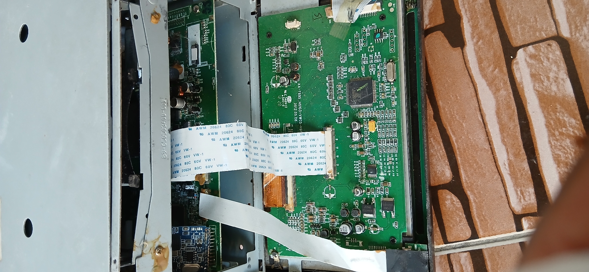 The image shows the internal components of an electronic device, likely a DVD player or similar media player, with its casing partially open. Two green circuit boards are visible, connected by white ribbon cables. One of the circuit boards has a large black integrated circuit chip in the center, surrounded by various smaller electronic components like resistors, capacitors, and transistors. The other circuit board, located at the top, appears to be a smaller daughterboard. The electronic components are mounted on a green printed circuit board (PCB). The device is placed on a surface that resembles a tiled floor with a brick pattern. There are no people or other subjects visible in the image. The lighting suggests an indoor environment. The image is a close-up view, focusing on the electronic circuitry. The provided crops highlight specific areas of the circuit boards and the device's casing. There is no information within the image that links it to the city of Samarinda, Indonesia.