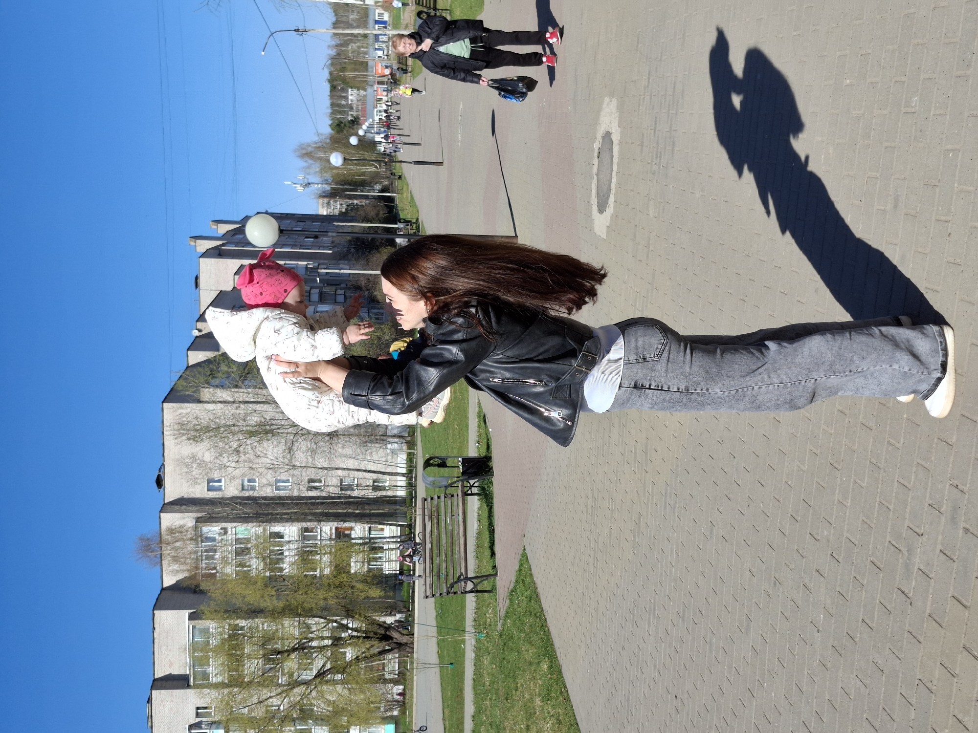 The image shows a sunny outdoor scene in Chaikovsky, Russia, on a paved walkway alongside a grassy area and park benches. A woman with long brown hair is holding a baby dressed in white with a pink hat. They are positioned so that the baby appears to be floating. In the background, there are apartment buildings and trees with budding leaves. Farther down the walkway, another person is walking. There is a long shadow cast on the pavement, likely from the woman and baby.