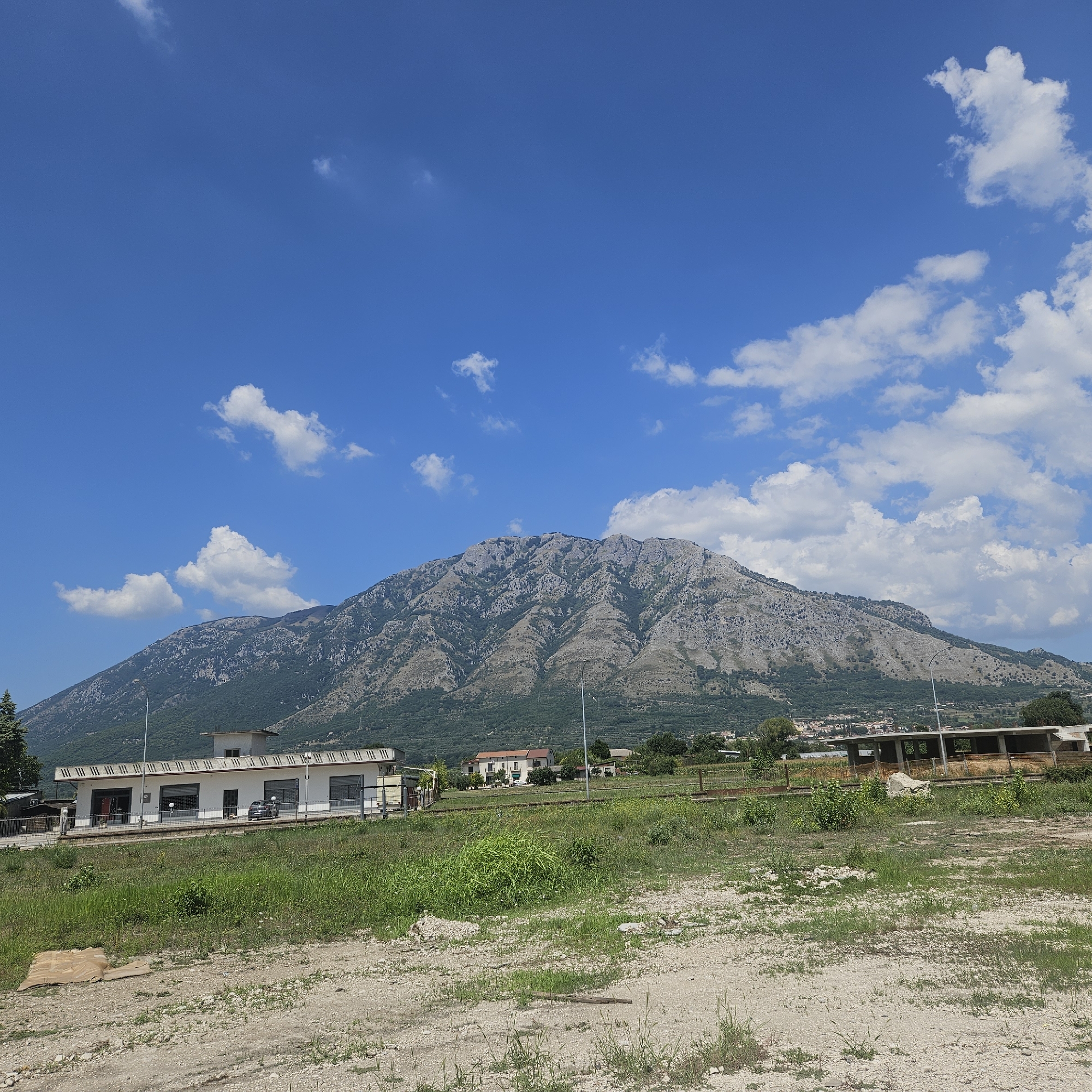 This is a landscape photograph taken on a clear day with a bright blue sky dotted with fluffy white clouds. In the foreground, a patch of dry, dusty ground with sparse vegetation is visible. Beyond this, a strip of green grass and some overgrown weeds are present. Further back, a white building with a long facade featuring several roller doors and windows is situated. A dark-colored car is parked in front of it. Behind this building, a residential area with houses and trees can be seen, set against the backdrop of a large, rugged mountain. The mountain is covered in grey rocks and sparse vegetation, with visible terracing or slopes. The overall scene depicts a rural or semi-rural environment with a prominent natural feature. The location is identified as Montesarchio, Italy.