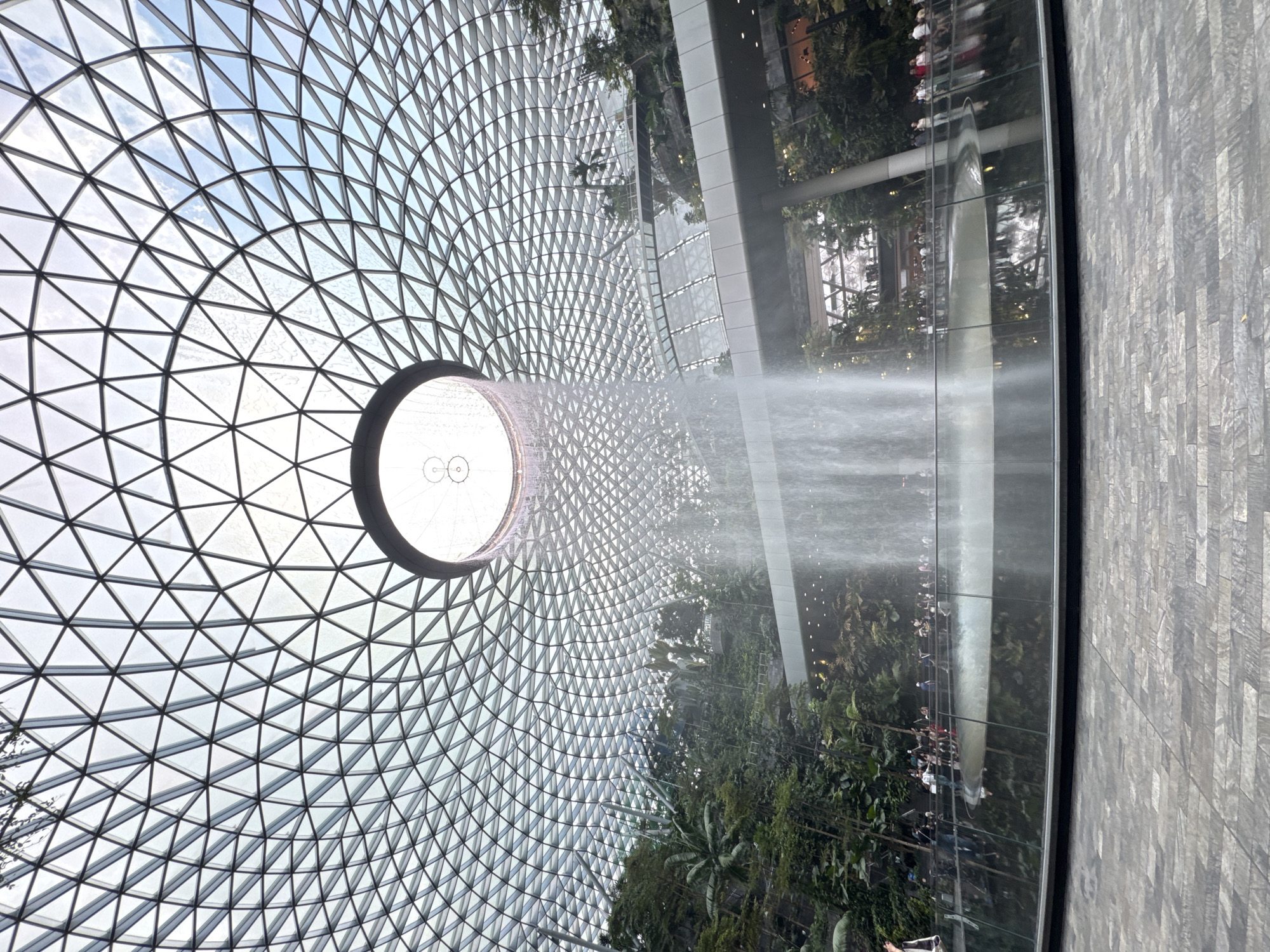 The image shows the Jewel Changi Airport in Singapore. A geometric glass dome structure dominates the view, with a circular opening in the center. Water is cascading down from the opening, creating a waterfall effect. Lush greenery and gardens are visible below the waterfall, and a wall with gray brick-like pattern extends on the right side of the image. The reflection of people, vegetation, and architectural elements can be seen on the reflective surface of the wall.