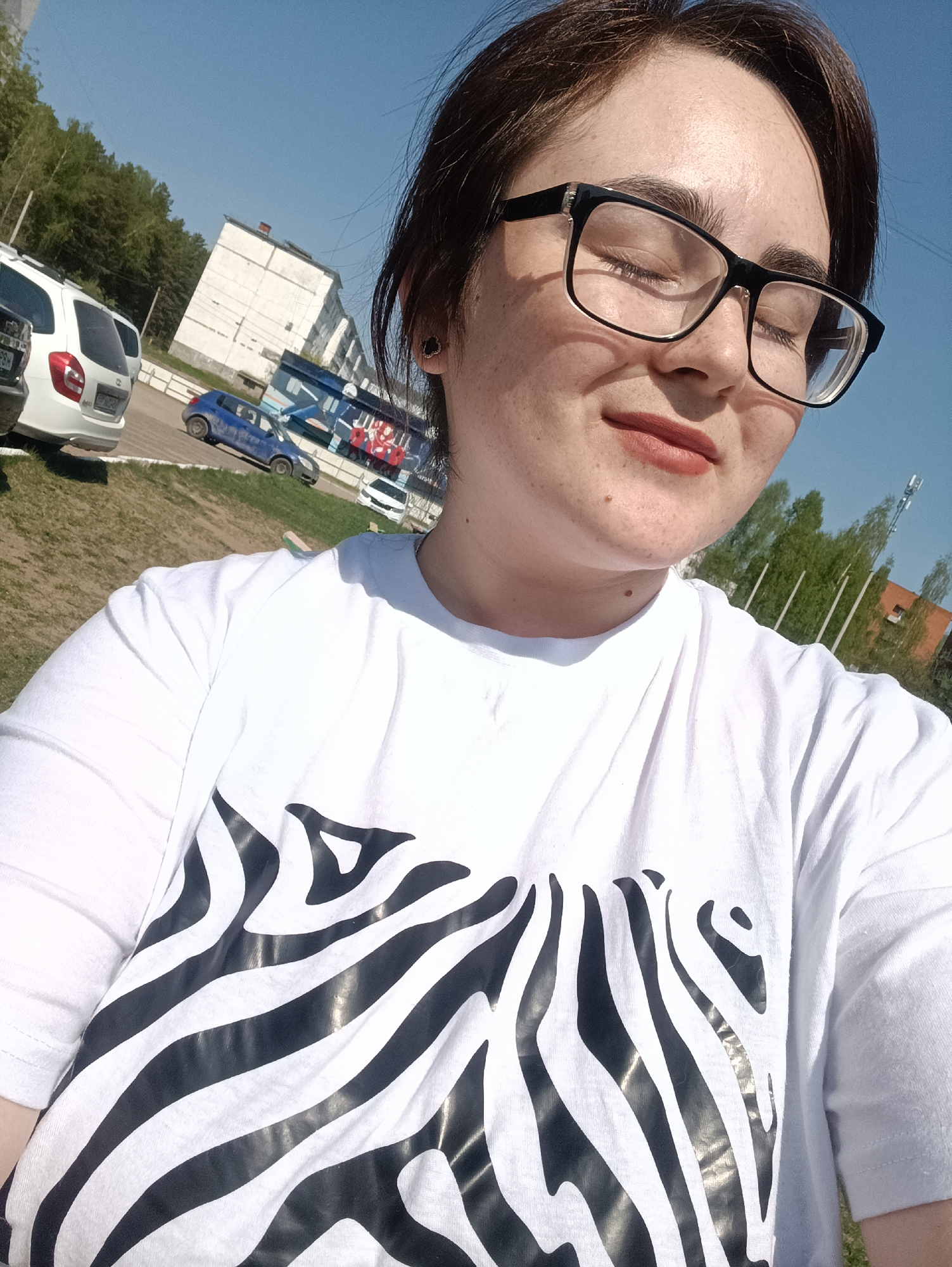A woman with brown hair and glasses is in the foreground, with her eyes closed and a peaceful expression. She is wearing a white t-shirt with a zebra-striped pattern. In the background, parked cars are visible, along with a multi-story building. The scene is outdoors on a sunny day. The location is the city of Noviy Urengoy, Russia.