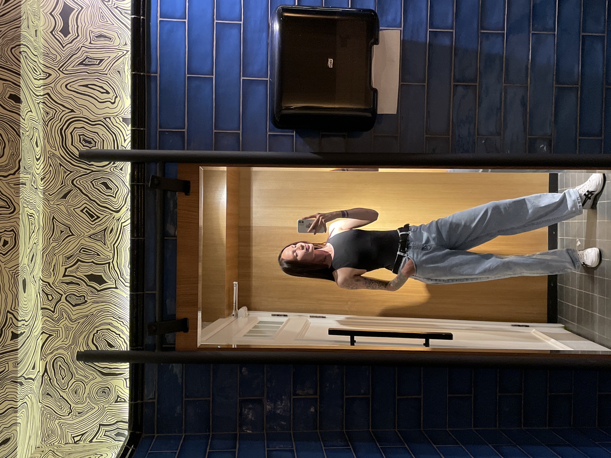 A woman is taking a mirror selfie in a restroom. She is wearing a black tank top, light blue jeans, a black belt, and white sneakers. She has a tattoo on her left arm. The restroom has blue tiled walls, a patterned wallpaper on the ceiling, and a wooden door. A black paper towel dispenser is mounted on the wall. The floor is tiled in a grid pattern. The scene appears to be an interior public restroom. No specific location within Izhevsk, Russia is identifiable from the image.