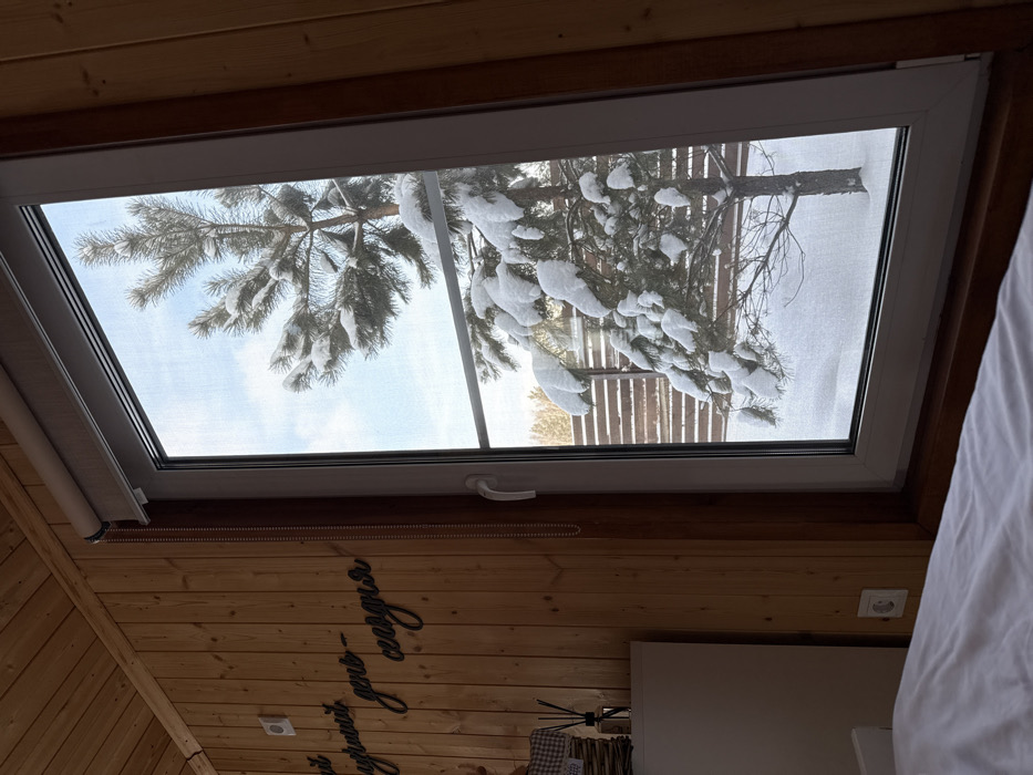 The image depicts an indoor scene looking out through a white UPVC-framed window to a snowy winter landscape in the city of Sysert, Russia. The interior features vertical light-toned wooden wall paneling on the right and upper left. A white-colored duvet on a bed occupies the lower foreground, extending from the left. An electrical outlet is visible on the wooden wall near the floor on the left. Further left, a white dresser or cabinet is partially visible, topped with a wicker basket and a reed diffuser containing dark liquid and reeds. Above this, on the wooden wall, black wooden letters spell out "Уютный день сегодня" (Cozy day today) in Cyrillic script. A beige roller blind is retracted at the top of the window, with a white plastic beaded pull chain hanging down the right side of the window frame.
Beyond the window, a coniferous tree, heavily covered in fresh white snow, stands prominently in the center. The ground is uniformly blanketed in snow. In the mid-ground, a horizontal slatted wooden fence runs across the scene. A clear, light blue sky is visible above the tree. No individuals or animals are present.
