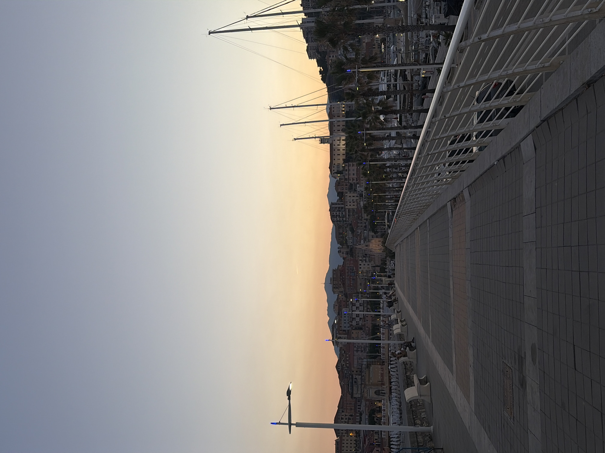 The image depicts a harbor scene at dusk in Imperia, Italy. The setting is a promenade with a tiled walkway and a white railing, leading the eye towards a cityscape nestled against a hillside. Tall sailboat masts dominate the upper left portion of the frame, silhouetted against a gradient sky transitioning from pale blue to a warm, peachy orange.
The cityscape is characterized by densely packed buildings with terracotta roofs, suggesting an Italian coastal town. Palm trees line the waterfront, and various boats are moored in the harbor, visible behind the promenade. People are seated on benches along the walkway, enjoying the evening atmosphere. Several streetlights with blue accents are visible, indicating the onset of night. The overall mood is tranquil and serene, with the warm colors of the sunset creating a peaceful ambiance. The time of day is clearly dusk, evidenced by the soft, diffused light and the transition of colors in the sky. The presence of sailboats and the architectural style of the buildings strongly suggest a Mediterranean coastal location, specifically identified as Imperia, Italy. There is no visible text in the image.