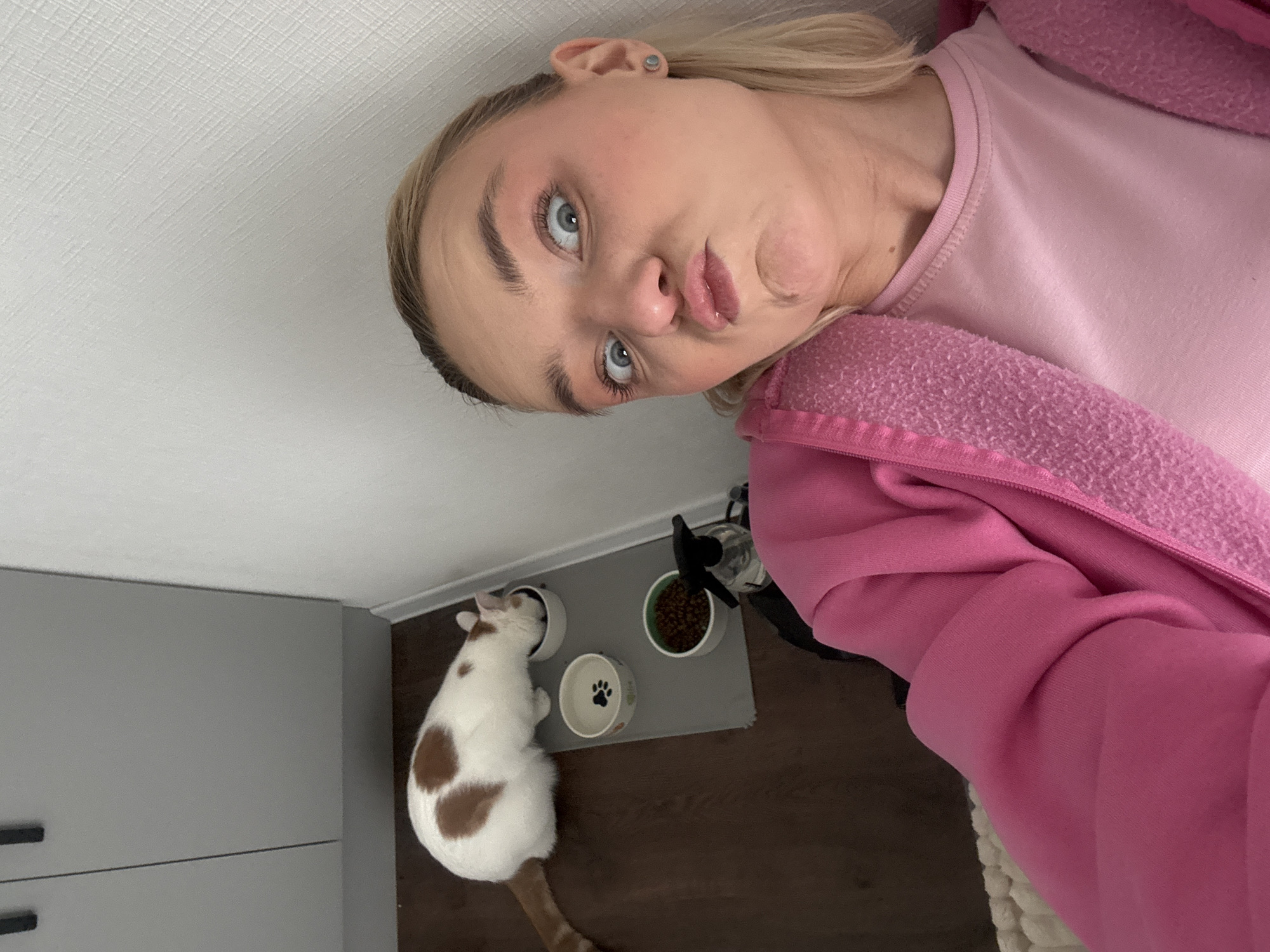 A selfie depicts a Caucasian woman indoors. She has blonde hair and blue eyes and wears a pink outfit. Below and to her left is a cat with white fur and brown spots eating from a bowl on a gray mat with two other bowls and a black object. The environment includes a gray cabinet, a white ceiling, and a dark floor. The location in Vsevolozhsk, Russia, is not identifiable from the image.