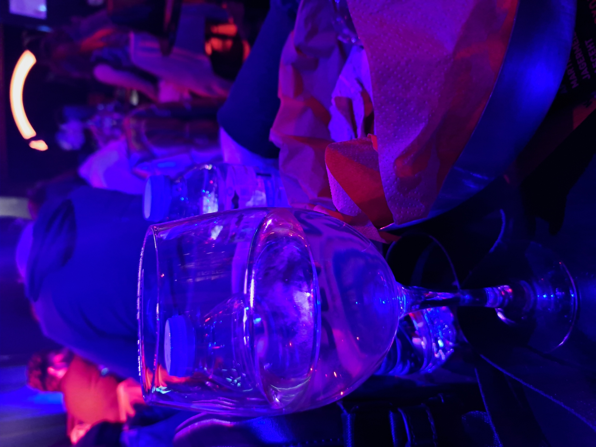 The image shows a wine glass lying on its side, illuminated by blue and purple lights. In the background, there is a metal bucket containing napkins with a red hue. The environment appears to be a bar or club due to the low lighting and presence of barware. The image was taken in Sofia, Bulgaria.