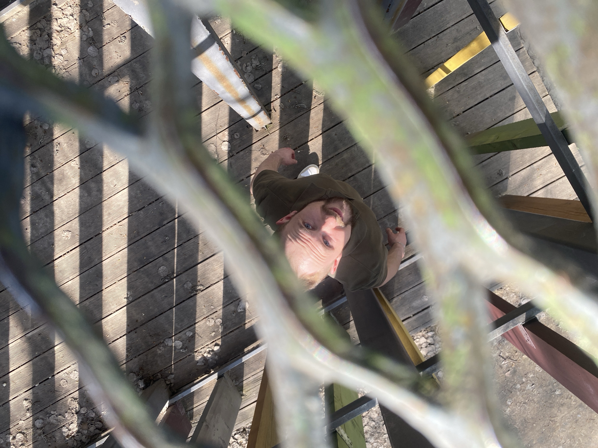 A man with fair skin, blue eyes, and short blonde hair is looking up at the camera from a platform made of wooden planks. The planks are horizontally aligned and covered in scattered debris, possibly leaves. A metal railing, partly obscured by the camera angle, surrounds the platform. The railing is painted in various colors, including green, yellow, and brown. The man is wearing a dark green shirt and white shoes. The photo appears to be taken from above, looking down through the railing. The overall lighting suggests a sunny day. The location is potentially within the city of Kazan, Russia, based on the request.