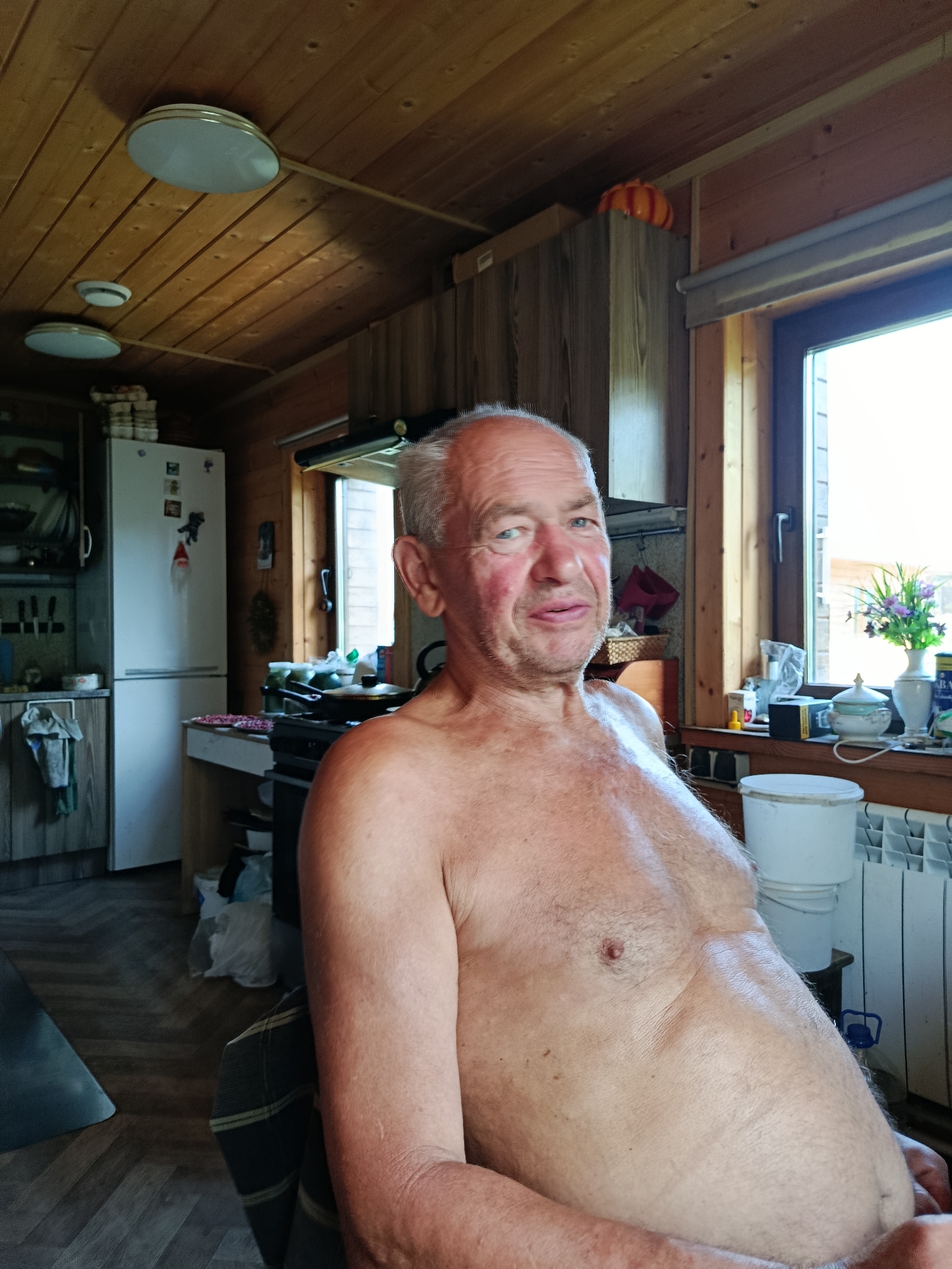 An outdoor medium shot shows a fair-skinned, middle-aged man with grey hair and light blue eyes, looking directly at the camera with a slight smile. He is shirtless, revealing his chest and abdomen. The man is sitting in an armchair, with his left arm resting on his lap and his right arm bent at the elbow. The background suggests an indoor domestic setting. The walls and ceiling are made of wood planks. To the left, a white refrigerator is visible, with several items on top. To the right, a window overlooks an outdoor scene, and a radiator is present. On a shelf by the window, there are several objects, including a vase with flowers and a white bucket. The man is in the process of sitting and looking forward. The lighting is bright, suggesting daytime.