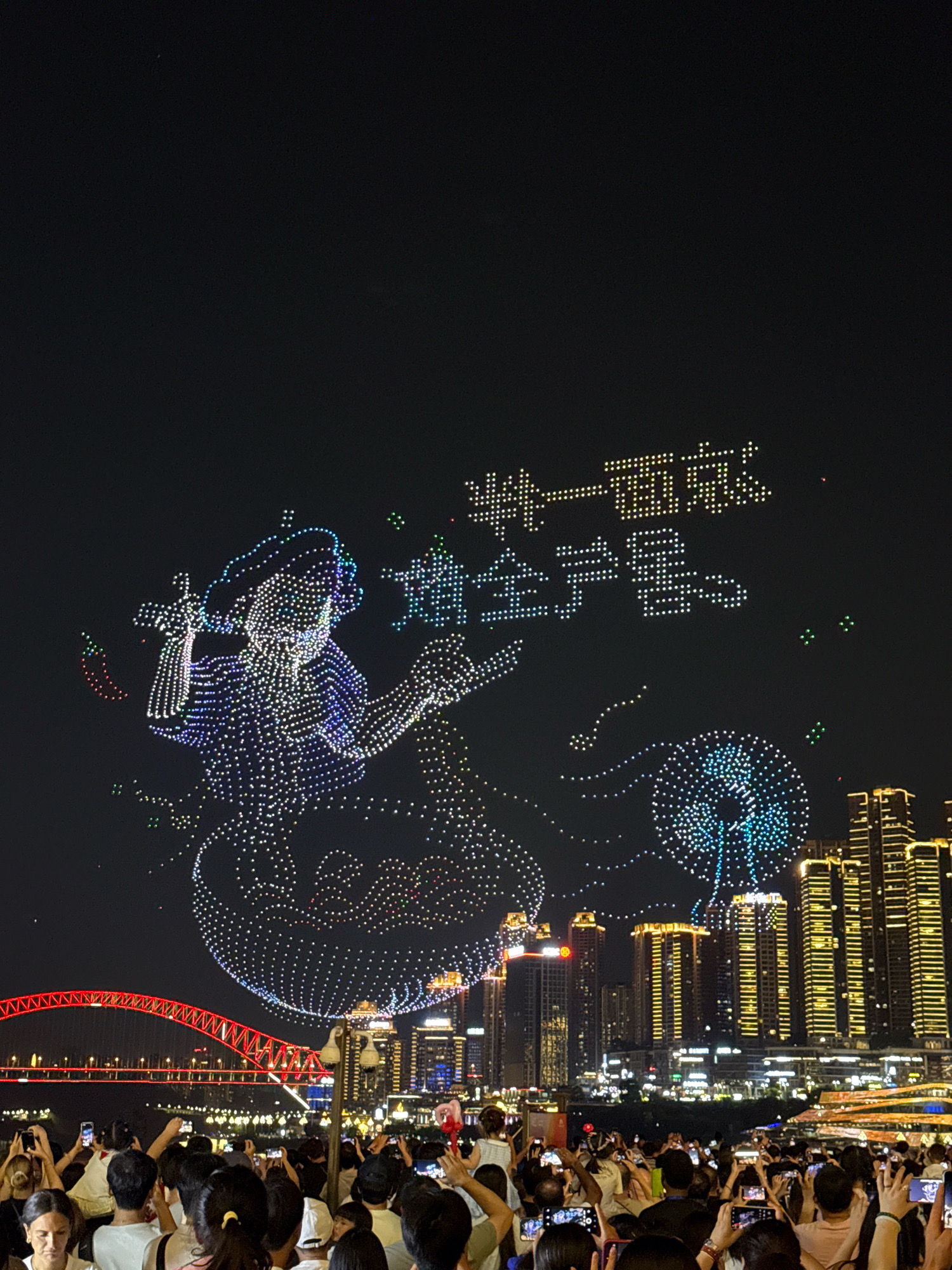 This image captures a nighttime drone show in the Yuzhong District of Chongqing, China. A large crowd of spectators is gathered on the waterfront, many holding up their phones to record the spectacle. The drone display features a formation of lights in the sky, including a detailed depiction of what appears to be a geisha or a similarly dressed figure, adorned with a wide-brimmed hat. To the right of the figure, Chinese characters formed by lights are visible, and further to the right, a stylized representation of a blooming flower or fireworks is illuminated. In the background, the illuminated skyline of Yuzhong District is visible, with tall buildings aglow with golden light. A red bridge arches across the water in the middle ground, further contributing to the vibrant urban night scene. The overall atmosphere is one of celebration and entertainment.