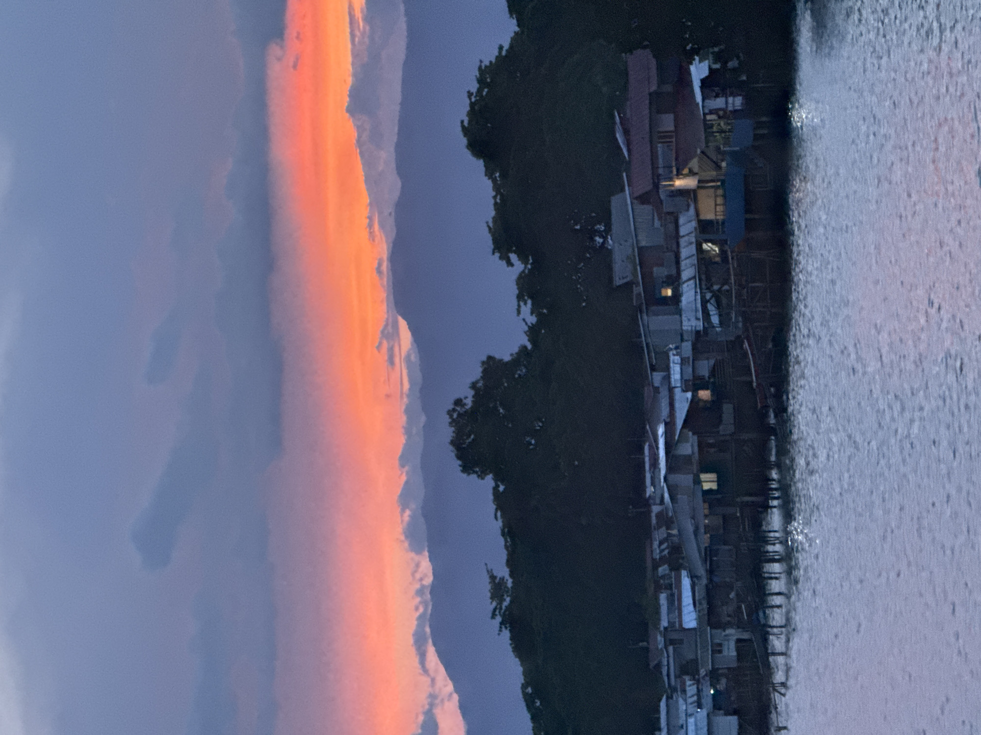 The image depicts a scene in Davao City, Philippines, likely near a waterfront area. There are shanties or houses built along the water's edge, supported by stilts or posts. The structures appear to be constructed from a variety of materials, including corrugated metal, wood, and other salvaged materials. The water is relatively calm and reflects the ambient light. In the background, there is dense foliage, suggesting a forested or heavily vegetated area behind the shanties. The sky is dramatic, with a mix of dark gray and orange clouds, indicating either sunrise or sunset. The lighting suggests that the time is dusk. There are no visible people or animals in the image. The image captures a mix of urban and natural elements, typical of waterfront communities in developing regions.
