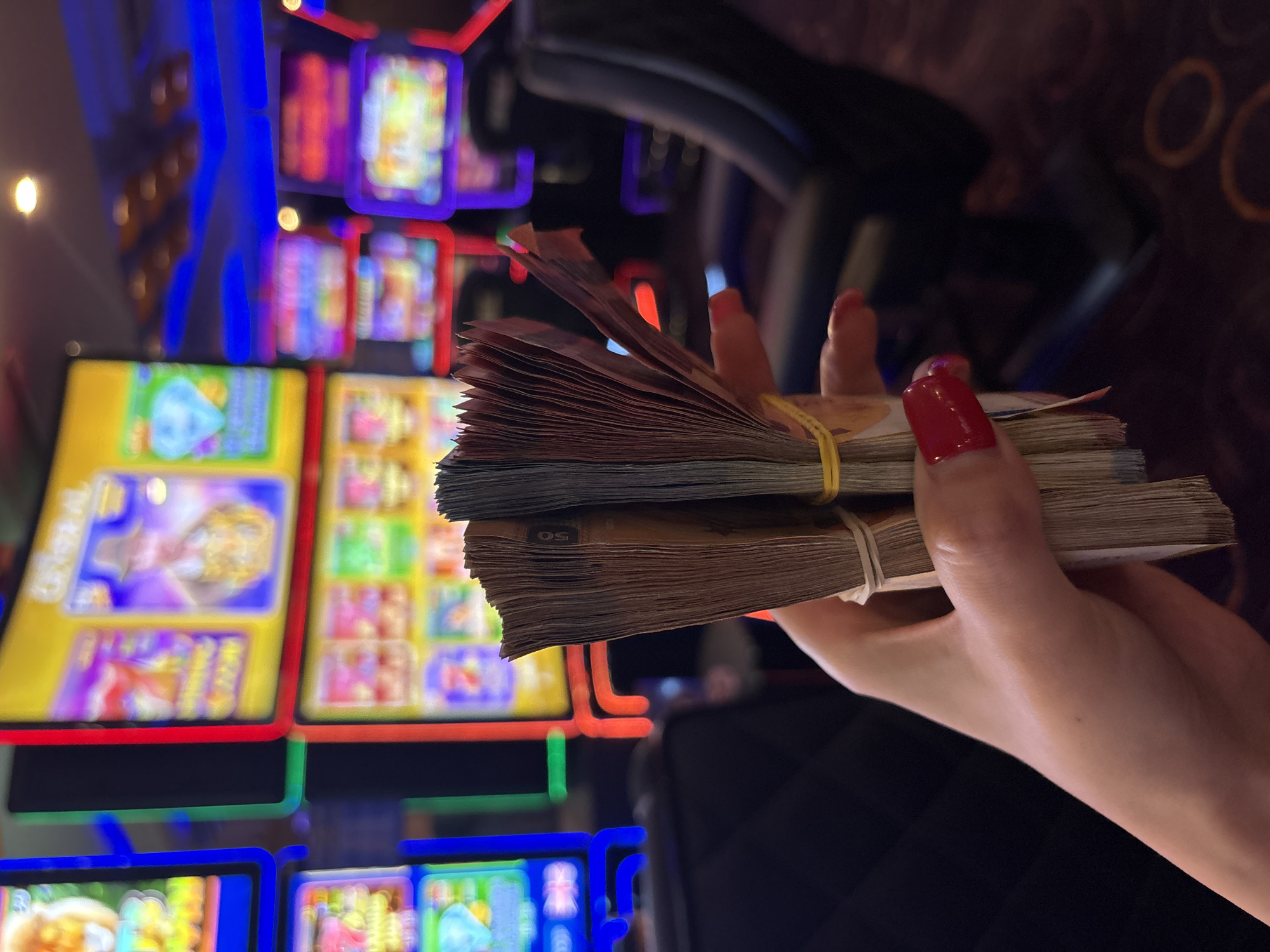 An angled, close-up shot features a person's hand holding several thick stacks of banknotes, secured with yellow rubber bands. The hand, adorned with prominent red polished fingernails, centers the bundles of currency in the foreground. Faintly visible on one of the banknote stacks is the number "50", suggesting a denomination.
The background is blurred but brightly lit, revealing the vibrant, multicolored screens of numerous slot machines or electronic gaming devices. These screens display abstract and colorful graphics, typical of casino games. The intense blue, red, and green artificial lights from the machines create a dynamic and energetic atmosphere, characteristic of a gambling establishment. A dark, upholstered chair or seat is partially visible in the upper right section of the frame.
The scene depicts a moment within what appears to be a casino or gaming hall, possibly located in Varna, Bulgaria. The individual is holding a substantial amount of cash, suggesting either recent winnings or funds prepared for gambling. The artificial lighting indicates that the time of day is not discernible. The image captures a static moment of possession within a lively, entertainment-focused environment.