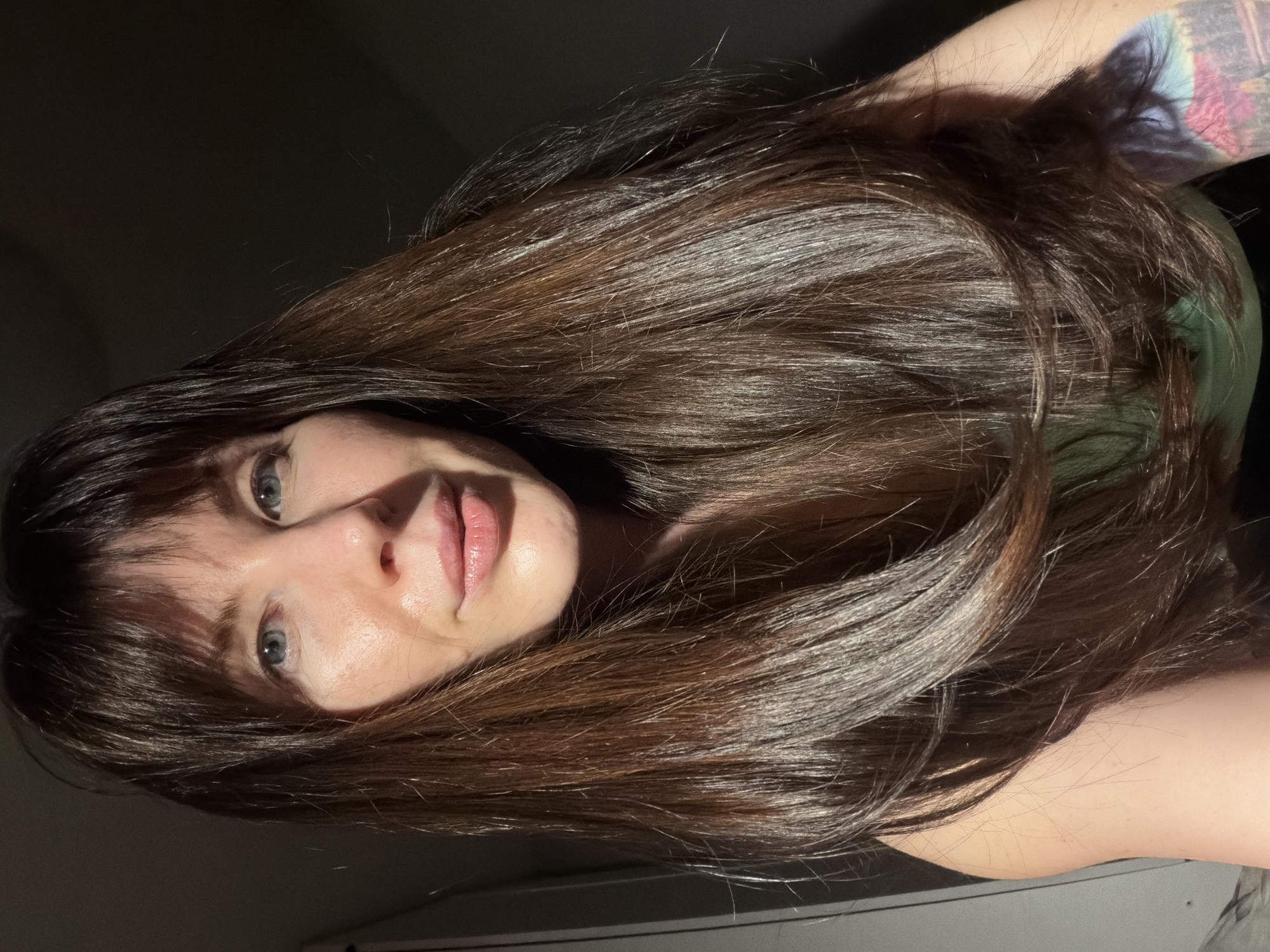 A woman with fair skin, blue eyes, and long brown hair is facing the camera. Her hair is partially covering her face, with strands across her forehead. She has pink lips and a small nose. Part of her right arm, with a visible tattoo, is in the upper right corner of the image. She is wearing a green top. The background is a dark, shadowed area. There is a white surface visible in the lower portion of the image. There is no evidence to suggest this image was taken in Perm, Russia.