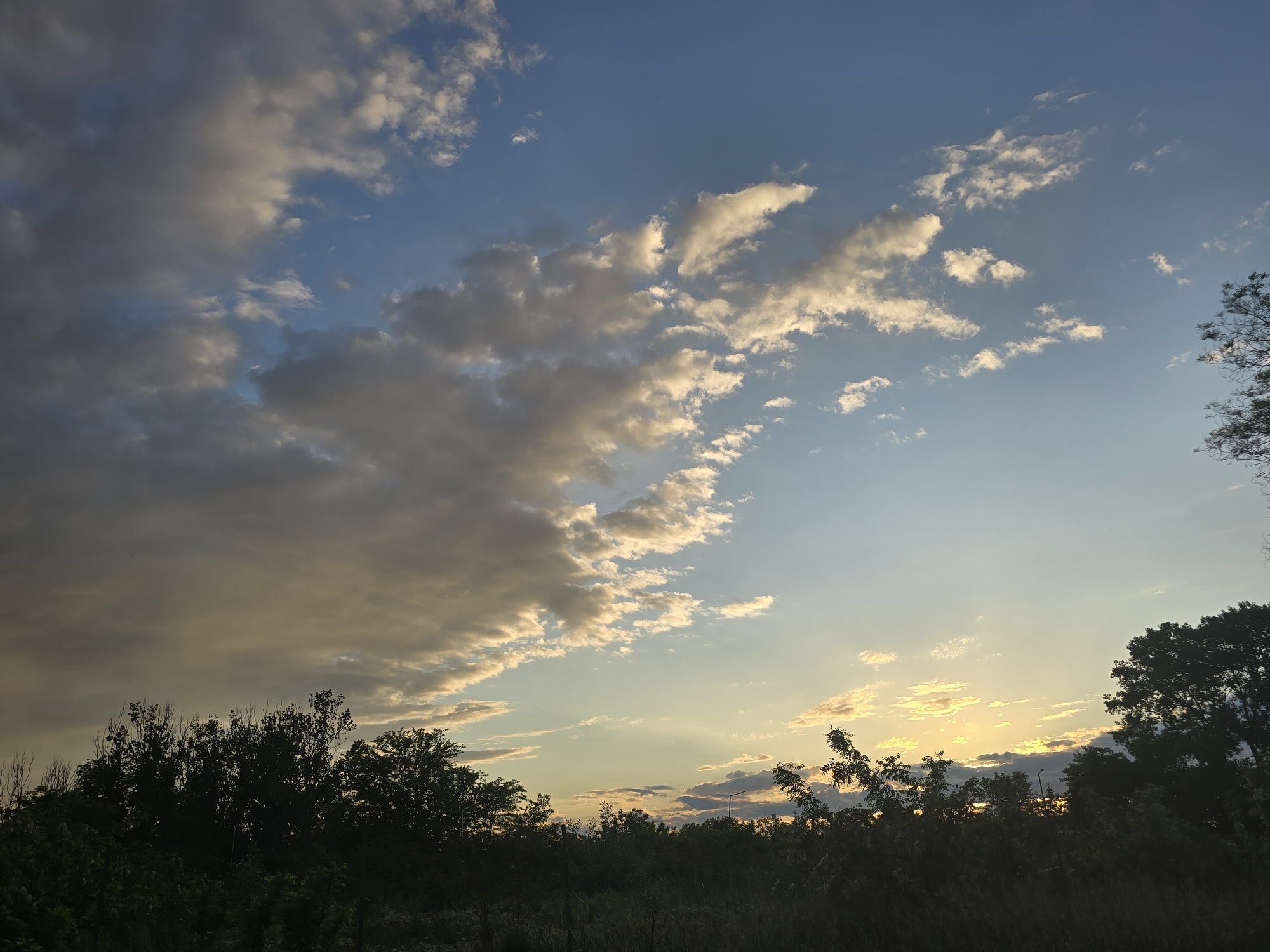 The image depicts an outdoor natural landscape captured during sunset or late afternoon. The sky dominates the upper two-thirds of the frame, showcasing a clear blue overhead that transitions to a lighter, golden-blue hue near the horizon. Scattered clouds vary in appearance; some are dense and grey, while others are illuminated by the setting sun, glowing with white and warm golden tones.
In the lower third of the image, the silhouettes of numerous trees and dense vegetation create a dark, undulating horizon line. These trees appear backlit, obscuring their details and emphasizing their dark forms against the brighter sky. A faint light pole is visible in silhouette against the lighter sky in the mid-right, positioned above the tree line.
There are no people or discernible activities taking place, presenting a serene and static scene. The notable details include the striking contrast between the dark foreground vegetation and the brightly lit sky and clouds, indicative of the golden hour. The overall impression is one of peaceful natural beauty as day transitions into evening. No visible text is present in the media file.