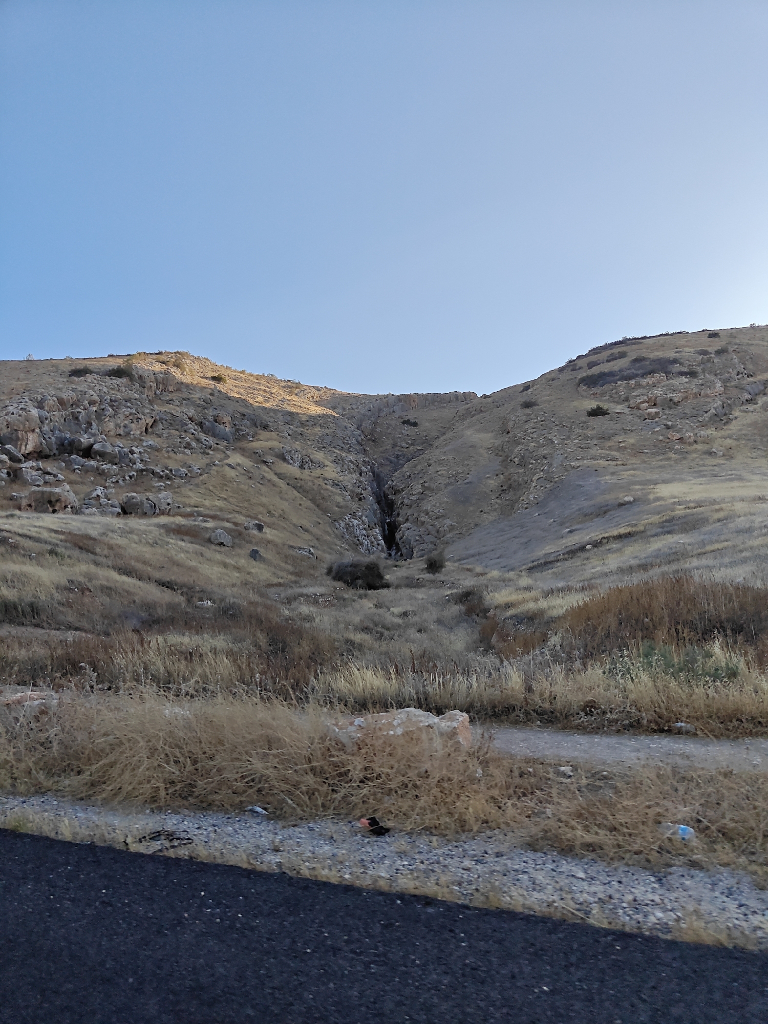 The image shows a desolate, rocky hillside under a clear blue sky. The terrain is arid, with sparse, dry grass and scrub. A deep ravine cuts into the hillside, appearing dark and shadowed within. Boulders are scattered across the slopes, and some appear to have fallen from higher up. In the foreground, a paved road runs along the bottom edge of the frame, with gravel and more dry vegetation bordering it. The overall impression is one of rugged, natural landscape. There are no people or discernible human activity visible in the scene, aside from the road. The location is the city of Al Jizah, Jordan.