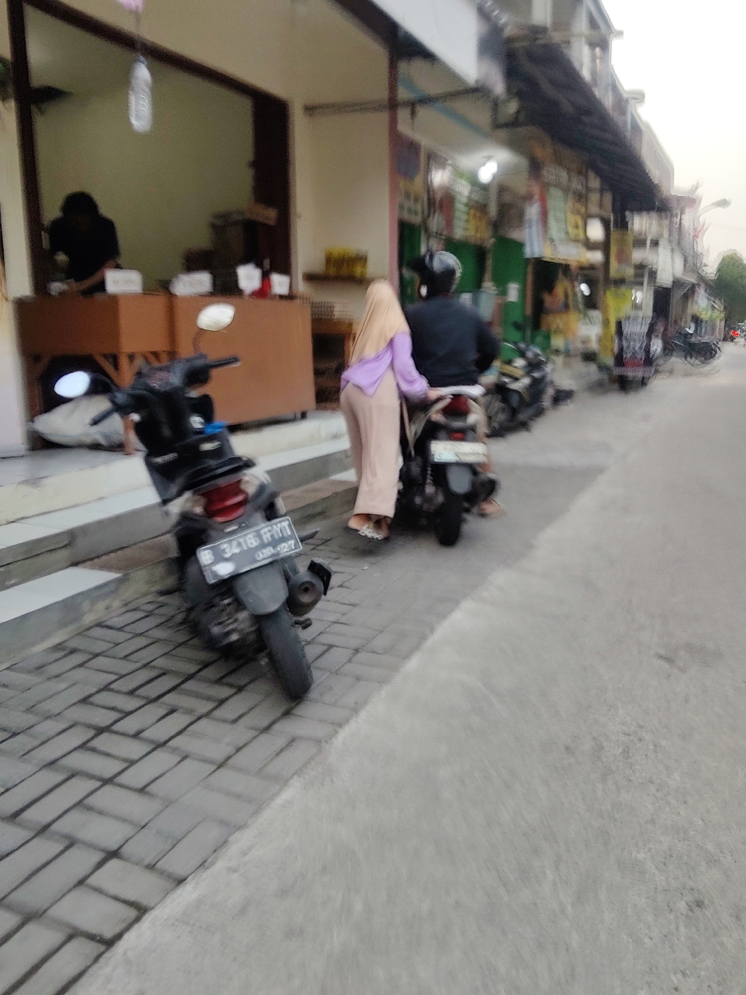 The image depicts a street scene in Mekarmukti, Indonesia, likely during daylight hours. In the foreground, a parked black scooter with a license plate that reads "B 3416 FFFT 09-27" is positioned on a gray brick sidewalk. To the right of the scooter, a person wearing a light purple hijab and a beige outfit is standing. Behind them, a person wearing a black helmet and a black shirt is seated on another scooter, also facing away from the camera. They are positioned in front of a building with a brown counter and a glass window, behind which a silhouette of a person in a dark shirt can be seen. The building appears to be a shop or food stall, with various items displayed inside. Further down the street, other shops and parked motorcycles are visible. The overall environment suggests a commercial or residential area with moderate foot and vehicle traffic. The lighting is somewhat diffused, indicating possible overcast conditions or late afternoon sun.