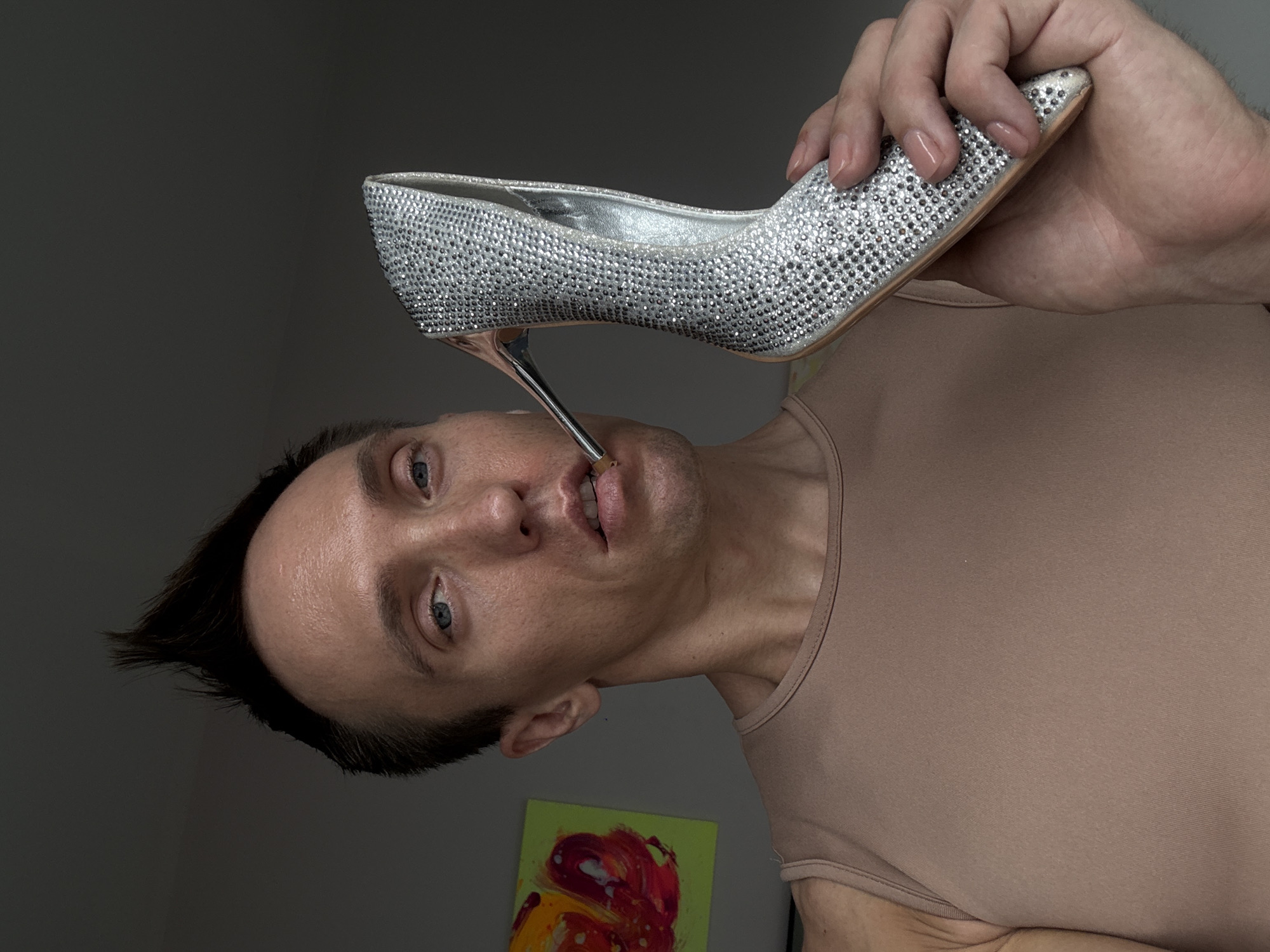 A close-up, waist-up, medium shot shows a fair-skinned person with short, spiky brown hair and blue eyes. They are wearing a tan, sleeveless top and holding a silver, rhinestone-covered high-heeled shoe by the heel with their right hand. The shoe's heel is positioned near their mouth, appearing as if it is being held between their lips. A small, abstract painting with red, yellow, and white colors on a green background is visible on the wall behind them to the left. The background is a plain, gray wall. The image appears to be taken indoors, with soft lighting. There is no indication of the location being in Baan Chaweng, Thailand.