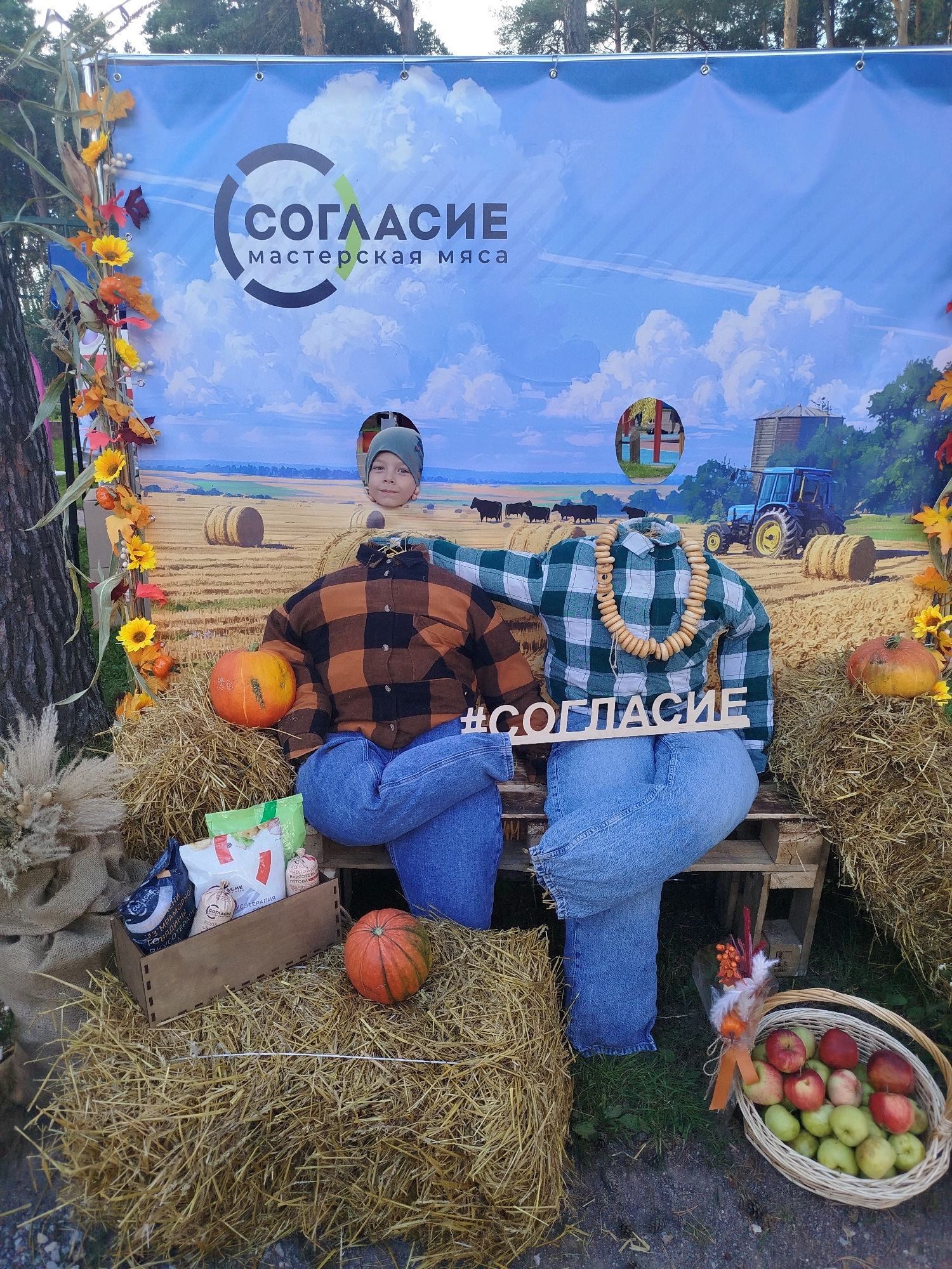 This is a photograph of an outdoor autumn-themed photo booth located in Zavodoukovsk, Russia. The backdrop depicts a rural farm scene with hay bales, a tractor, and cows under a blue sky with clouds. In the foreground, two headless mannequins are seated on a wooden bench, dressed in plaid shirts and jeans, positioned as if to be part of the photo booth. A young boy is visible peeking through a cutout in the backdrop, his head positioned where a head would normally be. Scattered around the mannequins are pumpkins, a basket of apples, bags of products, and decorative autumn foliage. The overall scene is designed for visitors to take humorous photos by inserting their faces through the cutouts in the backdrop.