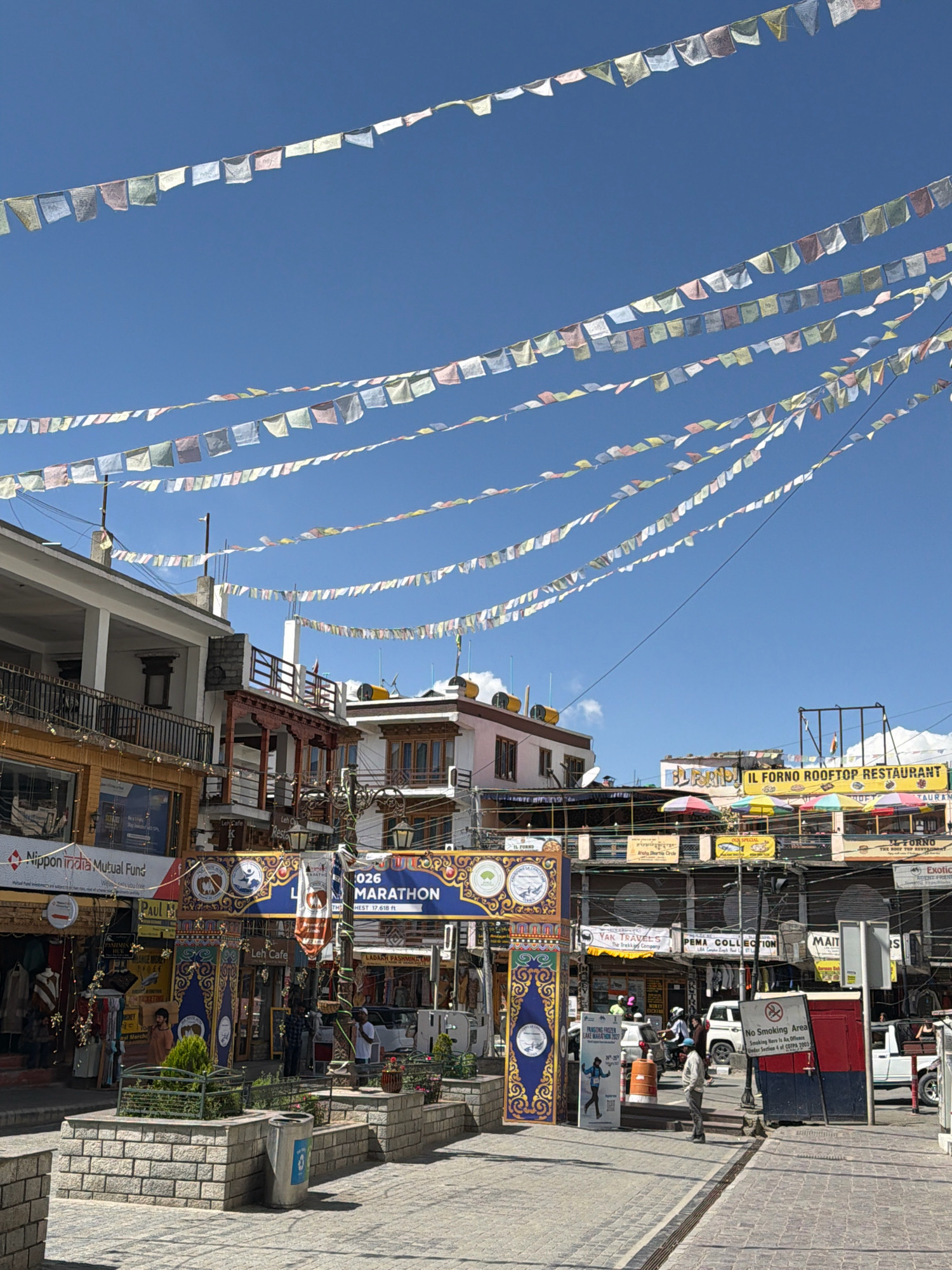 The image shows a street scene in Leh, India, on a clear day. Prayer flags are strung across the sky, creating a colorful canopy above the buildings. The buildings are a mix of traditional and modern architecture, with shops and restaurants on the ground floor and balconies on the upper levels.
In the foreground, there is a paved walkway with raised planters containing small trees and shrubs. A trash can is visible next to one of the planters. Several people are walking on the street and sidewalk. A white van is parked on the right side of the street, and a white SUV is visible behind it. A man is standing near a red and blue structure that appears to be a booth or kiosk. A "No Smoking Area" sign is posted on the structure.
Signs for various businesses are visible, including "Nippon India Mutual Fund," "Leh Cafe," "YAK TRAVELS," and "IL FORNO ROOFTOP RESTAURANT." A banner advertising the "PANGONG FROZEN LAKE MARATHON 2020" is also present. The overall atmosphere is that of a bustling street in a tourist town.