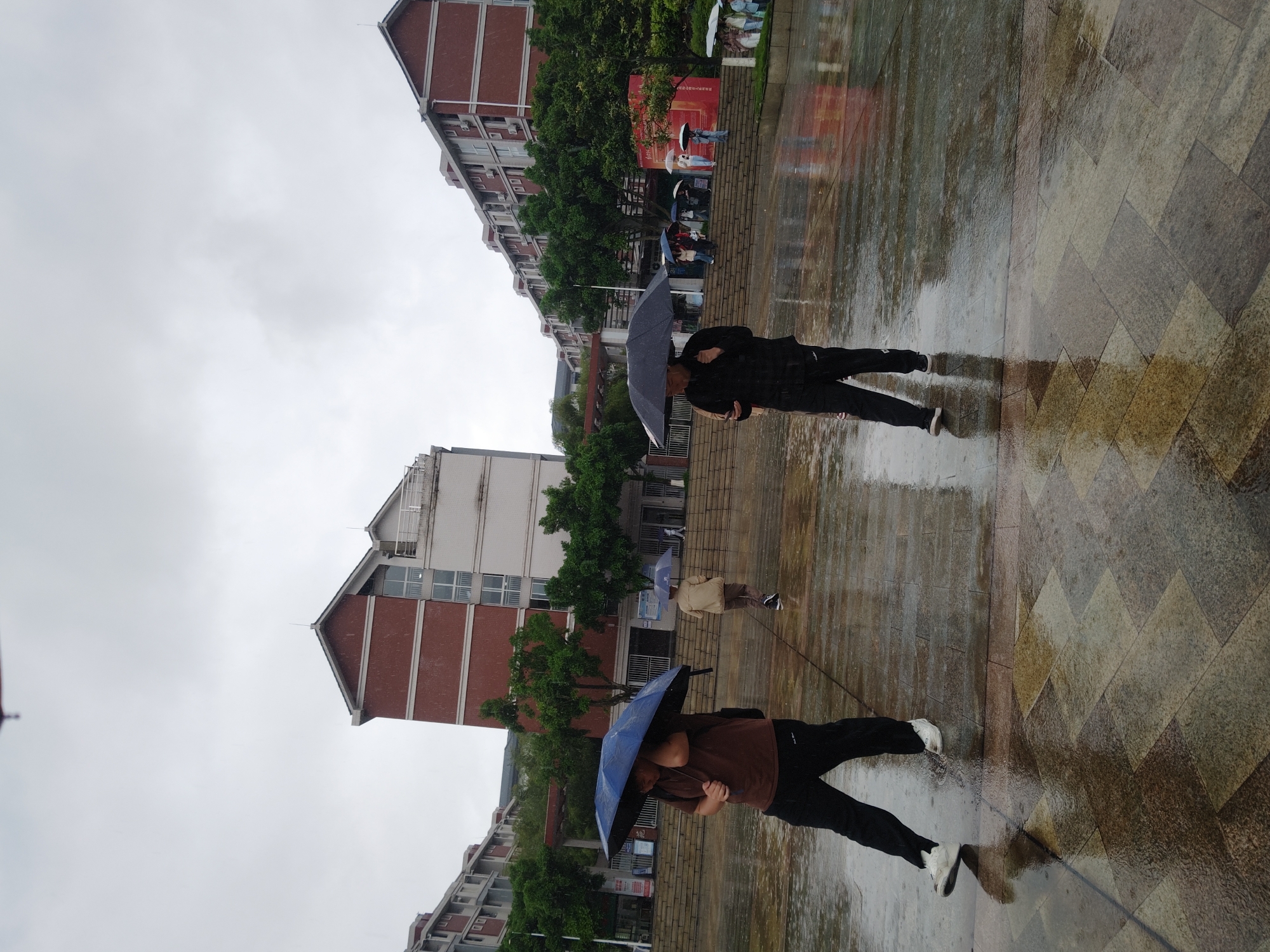 The image, taken in the Chongqing High-tech Zone, China, captures an outdoor scene on a visibly rainy or recently rained-upon day. The sky is overcast and uniformly grey, indicating heavy cloud cover. The ground is a paved area, likely a pedestrian walkway or public square, and is thoroughly wet, showing numerous puddles and strong reflections of the surroundings.
In the mid-ground, two individuals are walking away from the camera, each holding an open umbrella. One person wears dark clothing (a dark top and dark pants) and carries a dark grey umbrella. The other is dressed in a brown shirt and dark pants, shielding themselves with a blue umbrella. Both are walking on the wet, patterned pavement.
Further in the background, more pedestrians are visible, also holding umbrellas, suggesting ongoing rainfall. They are walking along a path bordered by green trees. Behind the trees, several multi-story buildings with red brick facades and white window frames are prominent, contributing to an urban or campus setting. The wet ground vividly reflects the buildings, trees, and the overcast sky, enhancing the sense of a rainy atmosphere. No discernible text is visible on any signs or banners. The scene depicts everyday activity continuing despite the wet weather conditions.