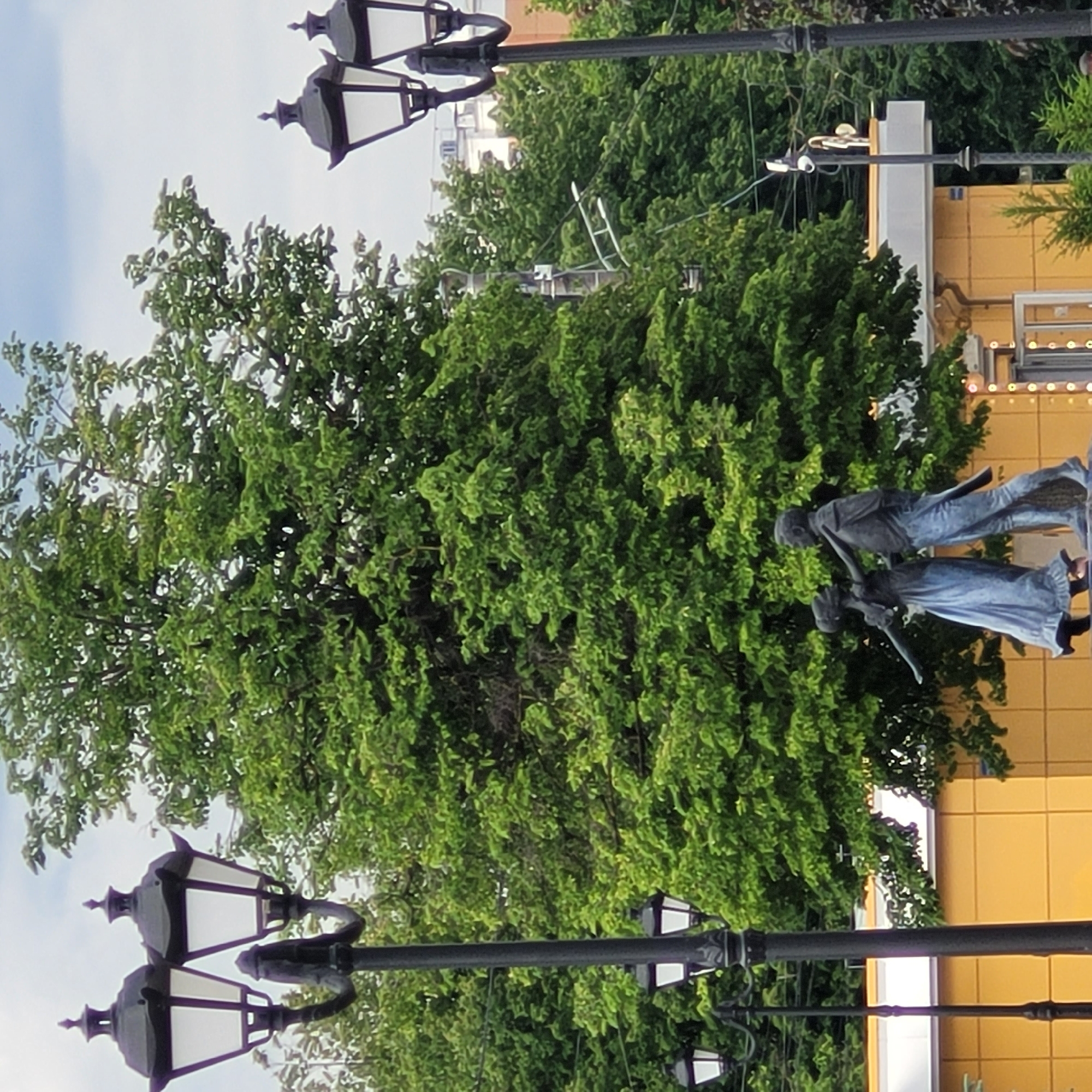 This media file captures an outdoor urban or park scene, presented with a vertical orientation (rotated 90 degrees clockwise).
The main subjects are a sculpture featuring two human-like figures, prominently placed towards the right side of the image, and a large, dense tree dominating the left and central portions. The male figure in the sculpture is dressed in a dark shirt and blue pants, seemingly lifting or holding the female figure, who wears a light blue dress with her arms extended. Their dynamic pose suggests dancing or a joyful interaction.
In the foreground, several ornate street lamps with black posts and white glass lanterns are visible; one is in the upper left corner and another is in the lower left, extending into the center. Behind the sculpture and tree, a building with a bright yellow facade is visible on the right, featuring decorative lights, possibly string lights, along its upper edge.
The scene is illuminated by bright daylight, with a clear or lightly clouded sky visible in the background, suggesting fair weather. The combination of the sculpture, mature trees, street lamps, and a building indicates an outdoor public space, such as a park, plaza, or city street. There is no visible text in the image.