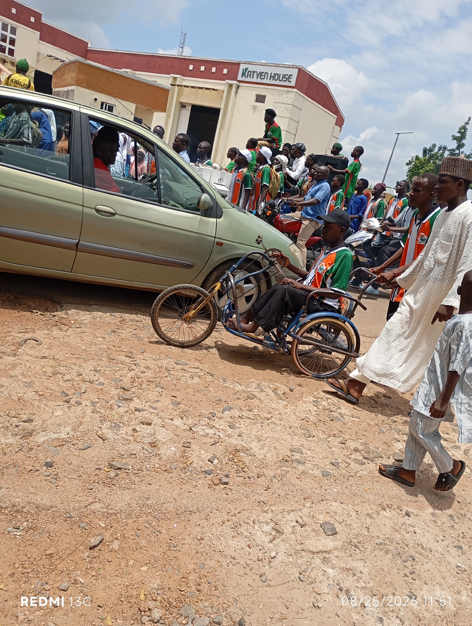 In Shendam, Nigeria, on a dusty road, a man in a wheelchair is being pushed by two other men. The man in the wheelchair is wearing a green and orange shirt and black pants. One of the men pushing the wheelchair is wearing a white robe and the other is wearing a blue shirt. A light green car is parked next to them. In the background, a building with the words "KATYEN HOUSE" is visible, along with a crowd of people, some of whom are wearing green and orange shirts. The sky is blue with some clouds. The ground is made up of dirt and gravel.