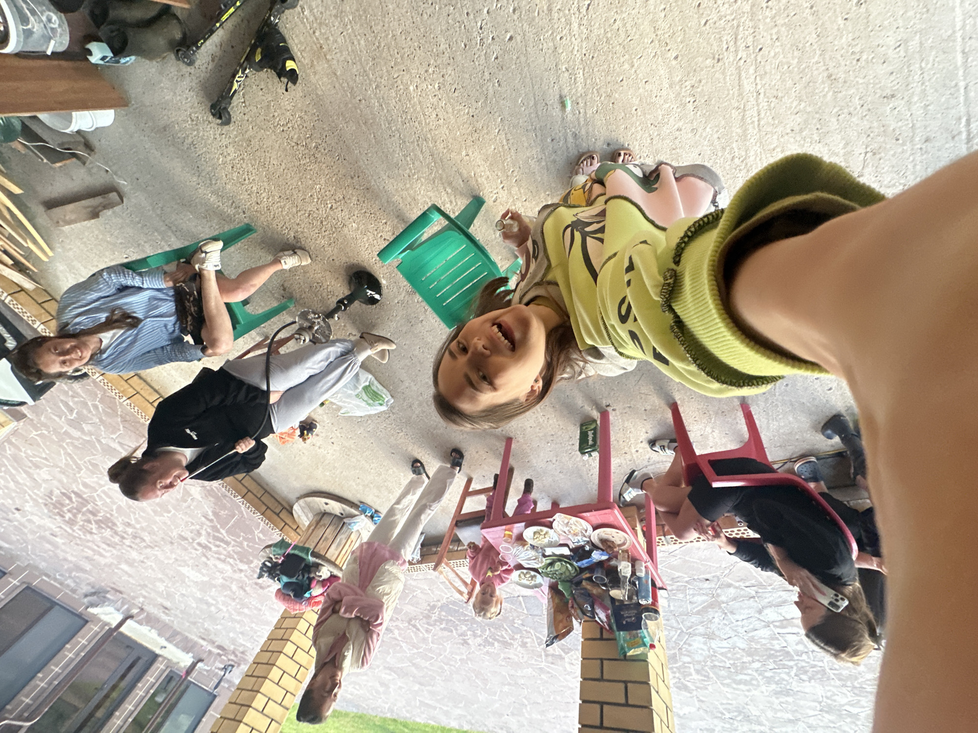 The image shows an outdoor scene at what appears to be a residential backyard or patio area. The ground is concrete, and there are brick elements visible on the left side, suggesting a building. There are several people present, including adults and a child. One woman is sitting on a green plastic chair, wearing a blue and white striped shirt and dark shorts, with her legs crossed. Another woman is standing, wearing a black jacket and grey pants, and holding a white object. A young child is sitting at a small red table covered with plates of food and various items. Another person is lying on a red chair, wearing black athletic shorts and a black long-sleeved shirt, with a phone in their hand. In the foreground, a person's arm and shoulder are visible, indicating the photo is taken from a first-person perspective. Several objects are scattered around, including skis, boots, a wooden spool, and plastic bags. The overall setting suggests a casual gathering or an informal outdoor activity. The image is taken in Пепеляева, Russia.