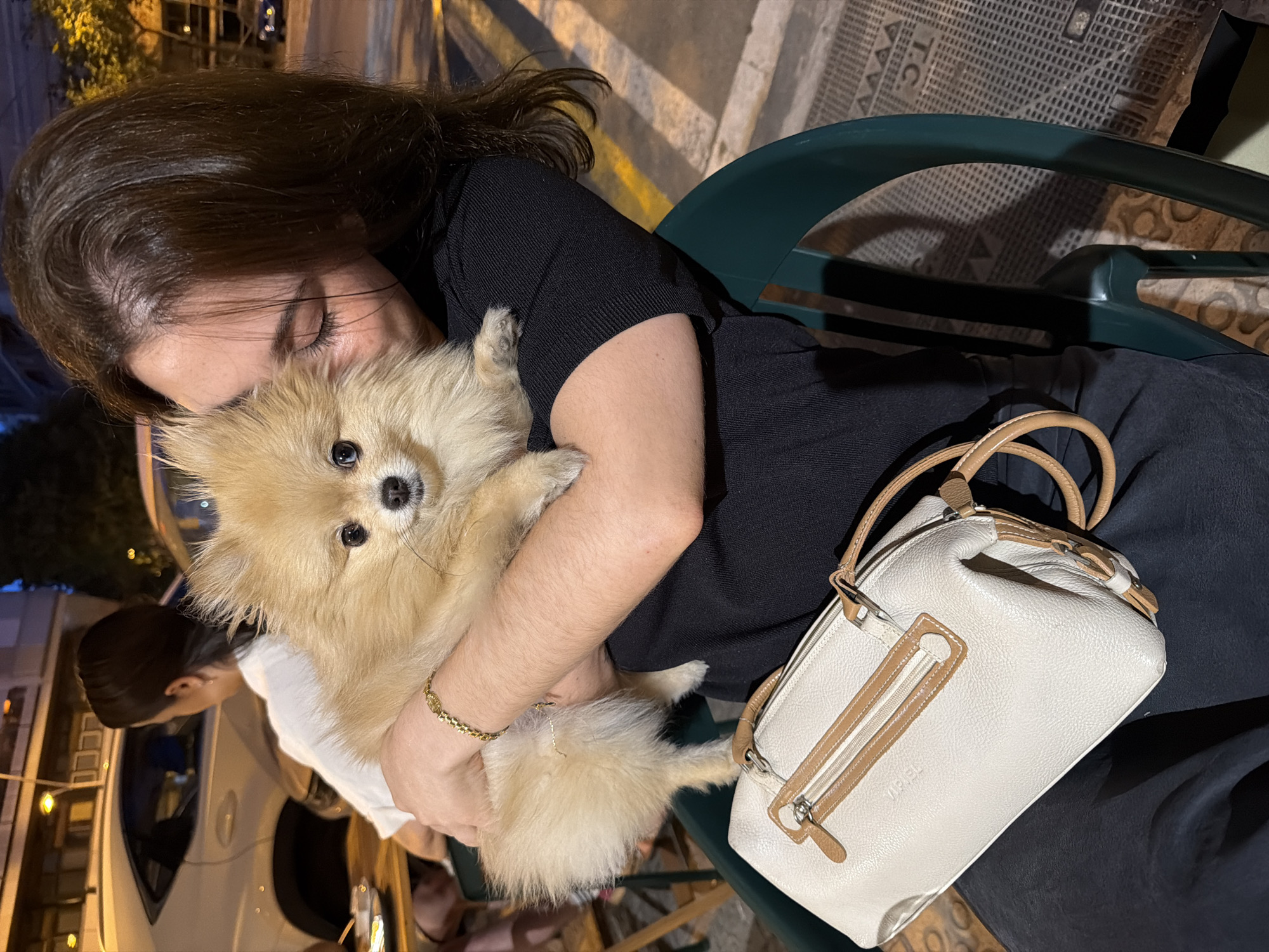 A young woman, with dark hair and fair skin, is holding a fluffy Pomeranian dog in her arms. She is wearing a black top and has a gold bracelet on her wrist. The dog is facing the camera, with its mouth slightly open. A cream-colored handbag with brown trim rests on her lap. The scene appears to be outdoors, possibly at a cafe or restaurant, with a green plastic chair to the right of the woman. In the background, there are cars and buildings, suggesting an urban environment. The lighting suggests it is evening or night. This image was taken in Barcelona, Spain.