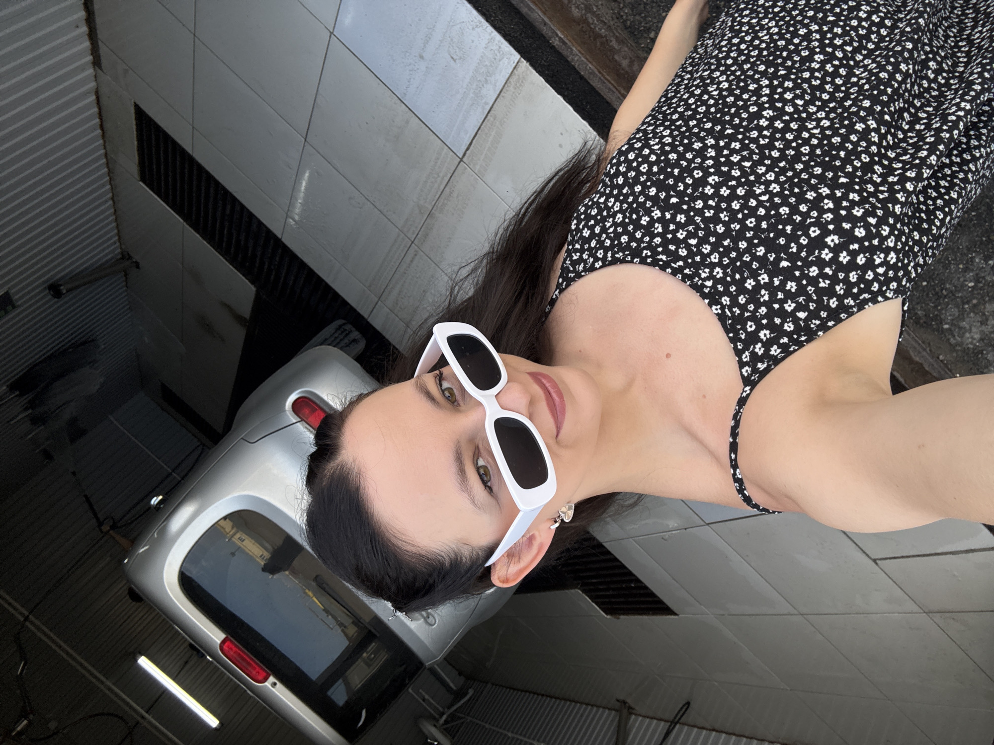 A woman is taking a selfie in a car wash. She is wearing a black dress with a floral pattern and white rectangular sunglasses. The car wash has a silver car in the background, tiled floors, and grates on the ground. The lighting suggests it is daytime. The location is identified as Chaikovsky, Russia.