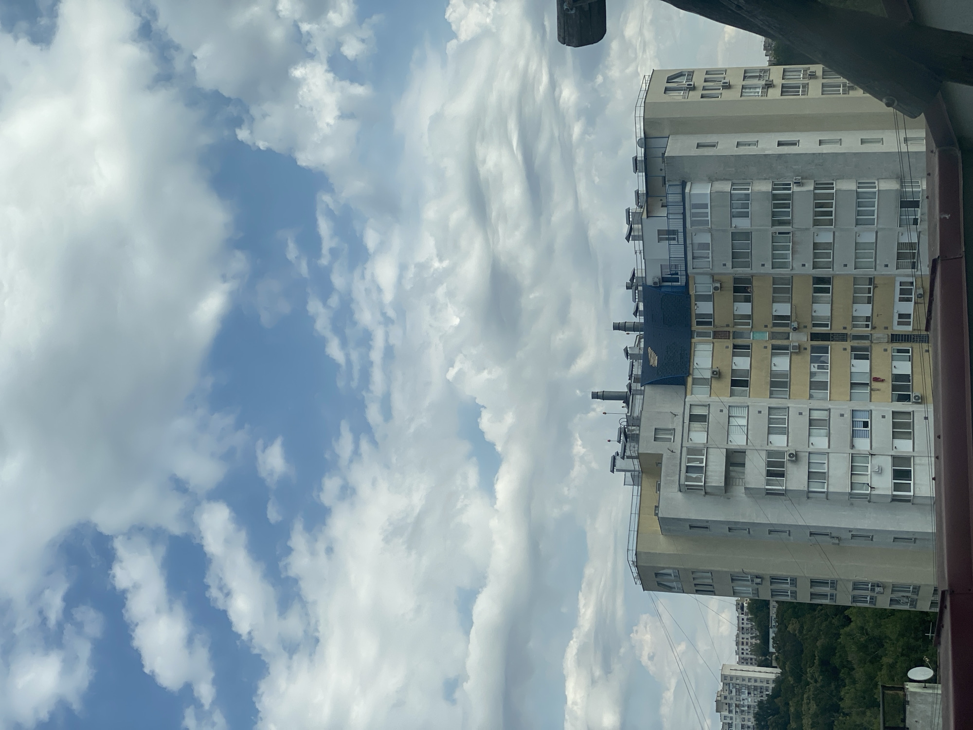 The image shows a multi-story residential building in Chișinău, Moldova. The building has a curved facade and is yellow and gray. It has numerous windows and balconies. On the roof, there are several chimneys and what appear to be solar panels. In the background, there are more buildings and a dense green forest. The sky is partly cloudy with a mixture of blue sky and white clouds. In the foreground, there is a dark, blurry object, possibly a piece of roofing or a piece of equipment, obscuring the lower right corner of the image. There are no people visible in the image.