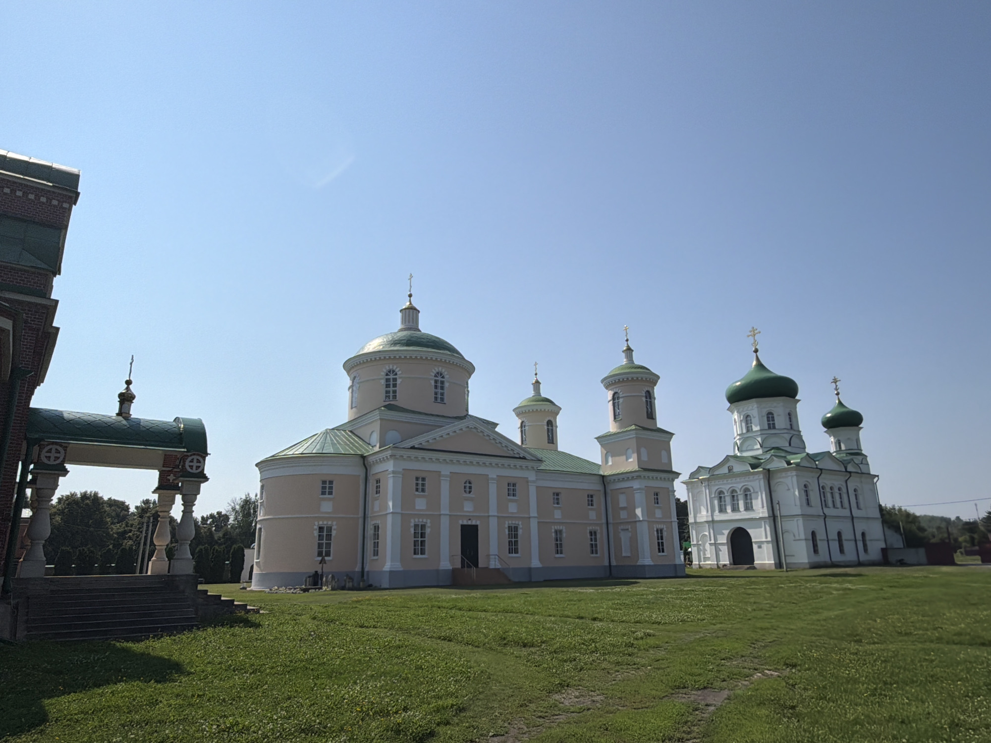 The image depicts a sunny daytime scene featuring several religious buildings in Troekurovo, Russia. In the foreground, a meticulously mowed green lawn stretches towards the structures. To the left, a portion of a red brick building with a green roof is visible, featuring a covered entranceway with ornate pillars and steps leading up. Further into the scene, a large, pale yellow building with a green domed roof and multiple windows stands prominently. To its right, a white building with a prominent green dome and several smaller domes, also topped with crosses, is situated. The sky is a clear, bright blue. No people are visible in the frame. The environment suggests a well-maintained religious complex or monastery.