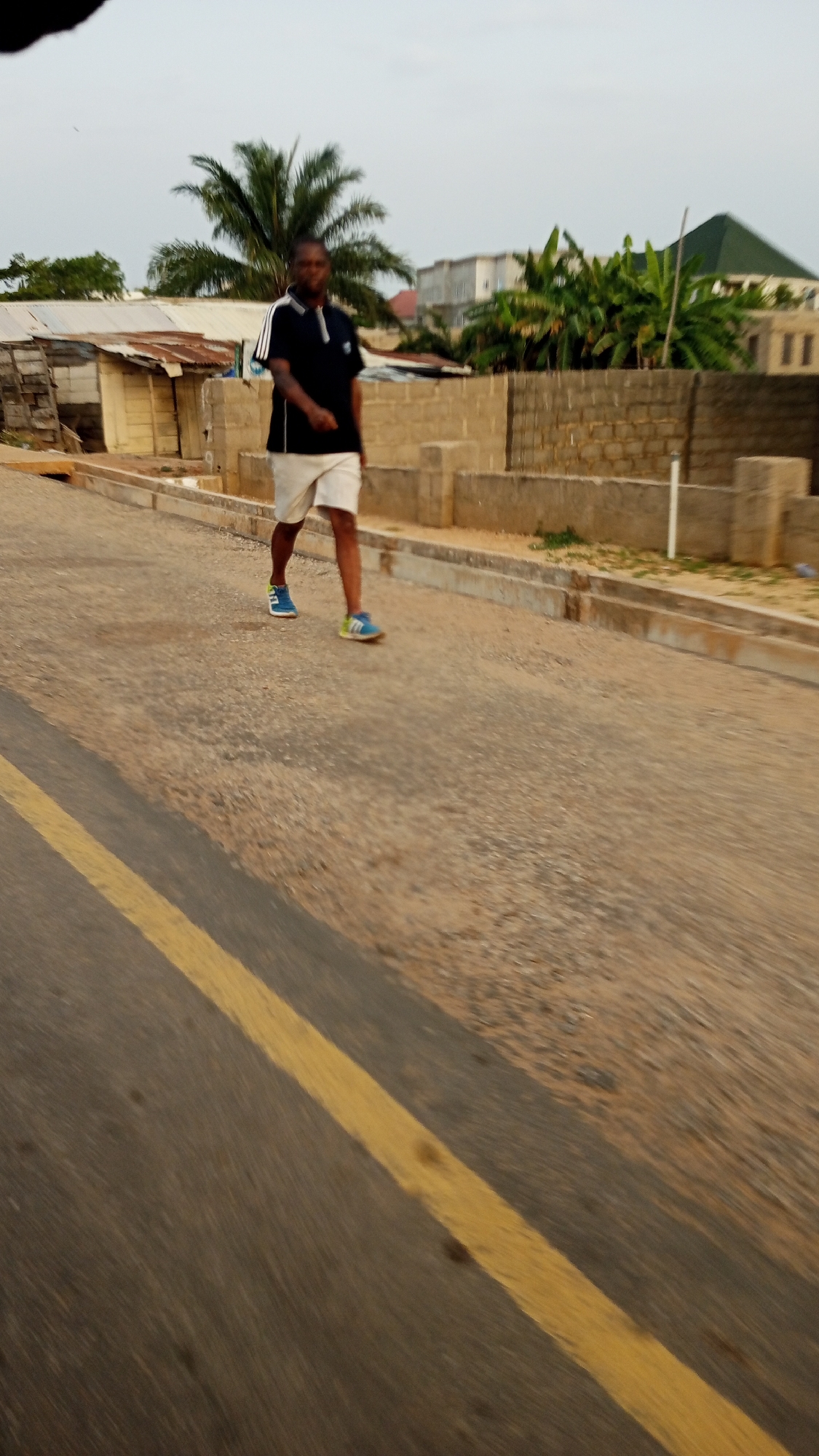 A man is walking on a street in an urban area. The street has a yellow painted line running along the left side of the road. In the background, there are a number of buildings. There is a palm tree behind the man.