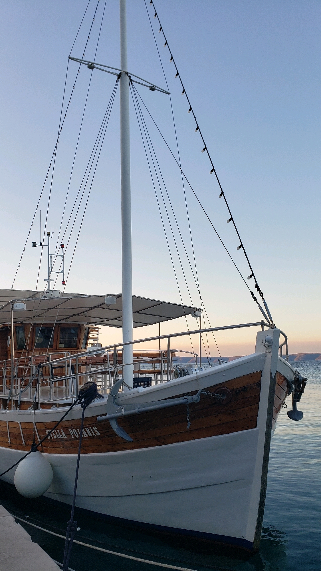 The image shows a docked boat named "STELLA POLARIS" with a white hull and wooden accents. The boat has a mast with numerous wires and a string of lights. A white fender is attached to the side of the boat. The boat is moored in calm water, with a pier visible in the foreground. In the background, a coastline with hills can be seen under a clear sky with a hint of sunset colors. The scene suggests a serene maritime setting, possibly in Općina Tučepi, Croatia, given the characteristic Adriatic coastline.